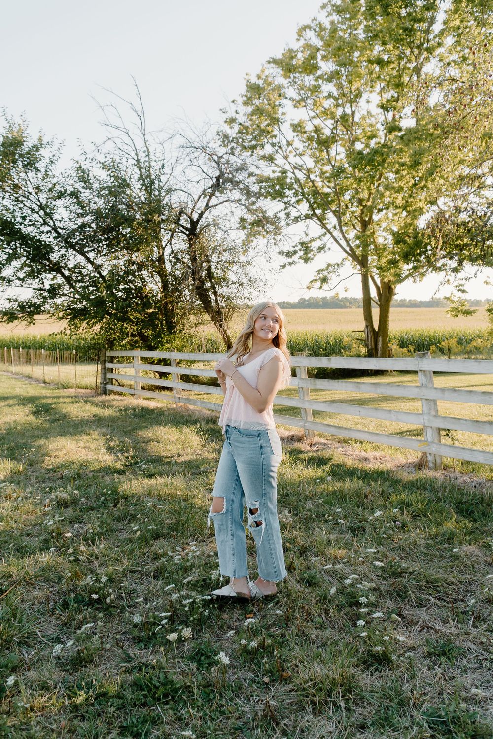 Person in light pink top and distressed jeans poses by white wooden fence in sunny rural setting at golden hour.
