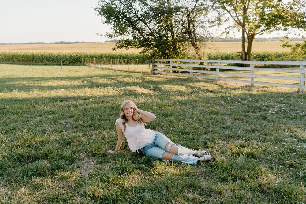 Person relaxing in a grassy field at sunset with a white fence and cornfield in the background.