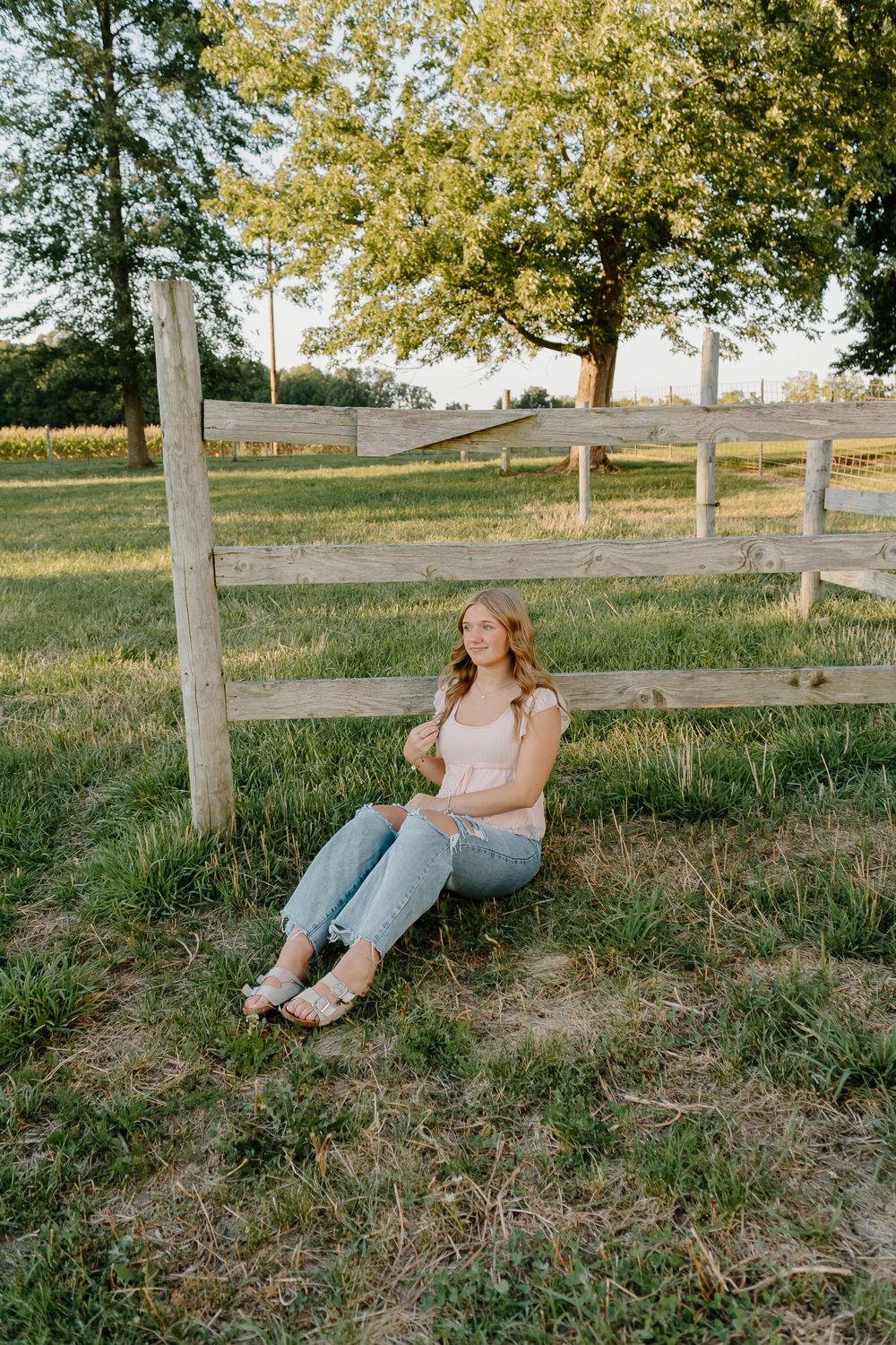 A person in casual attire sits on a grassy slope by a wooden fence during golden hour.
