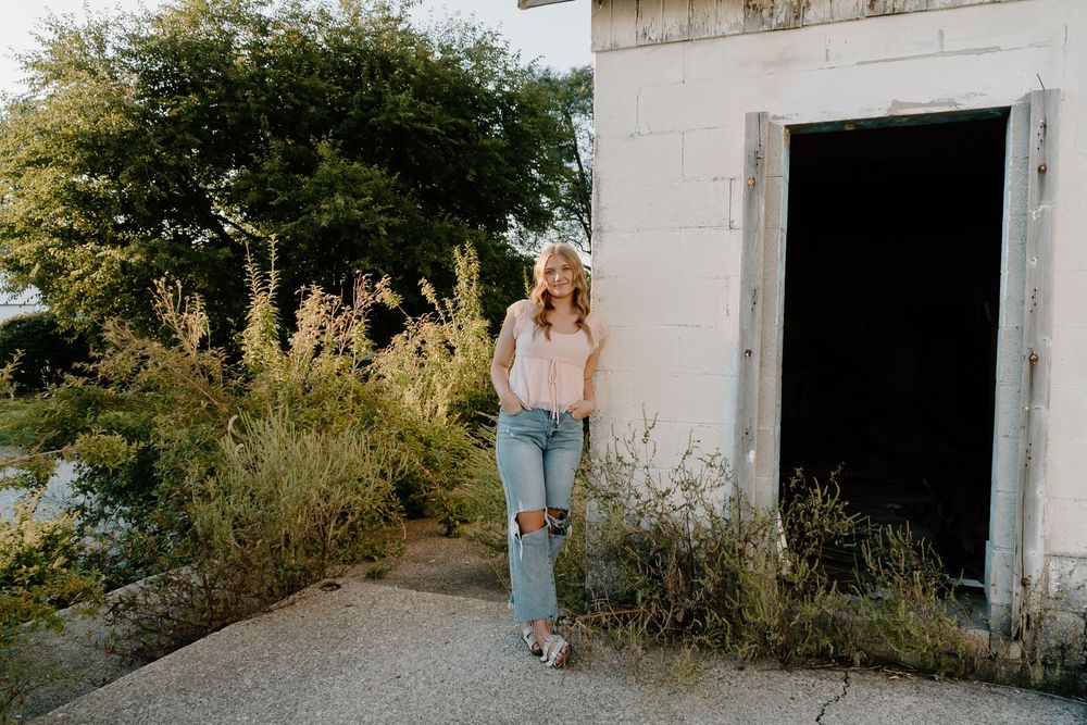Someone in ripped jeans and a white top stands near a rustic white building entrance surrounded by wild plants.