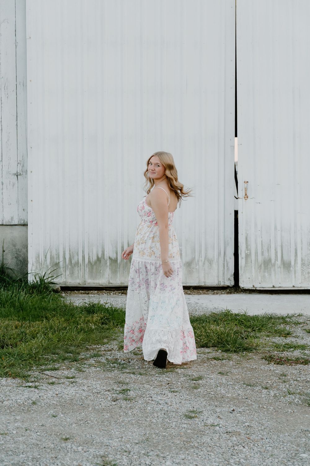 A child in a white lace dress twirls and plays in front of a white barn wall on a gravel surface.