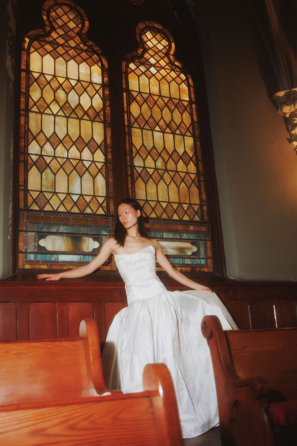 A white dress catches sunlight through stained glass church windows as someone poses on wooden pews.