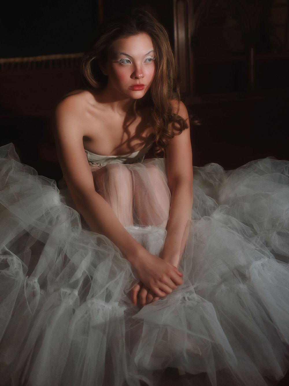 A model in a white tulle ballet-style dress poses dramatically in moody dark lighting.