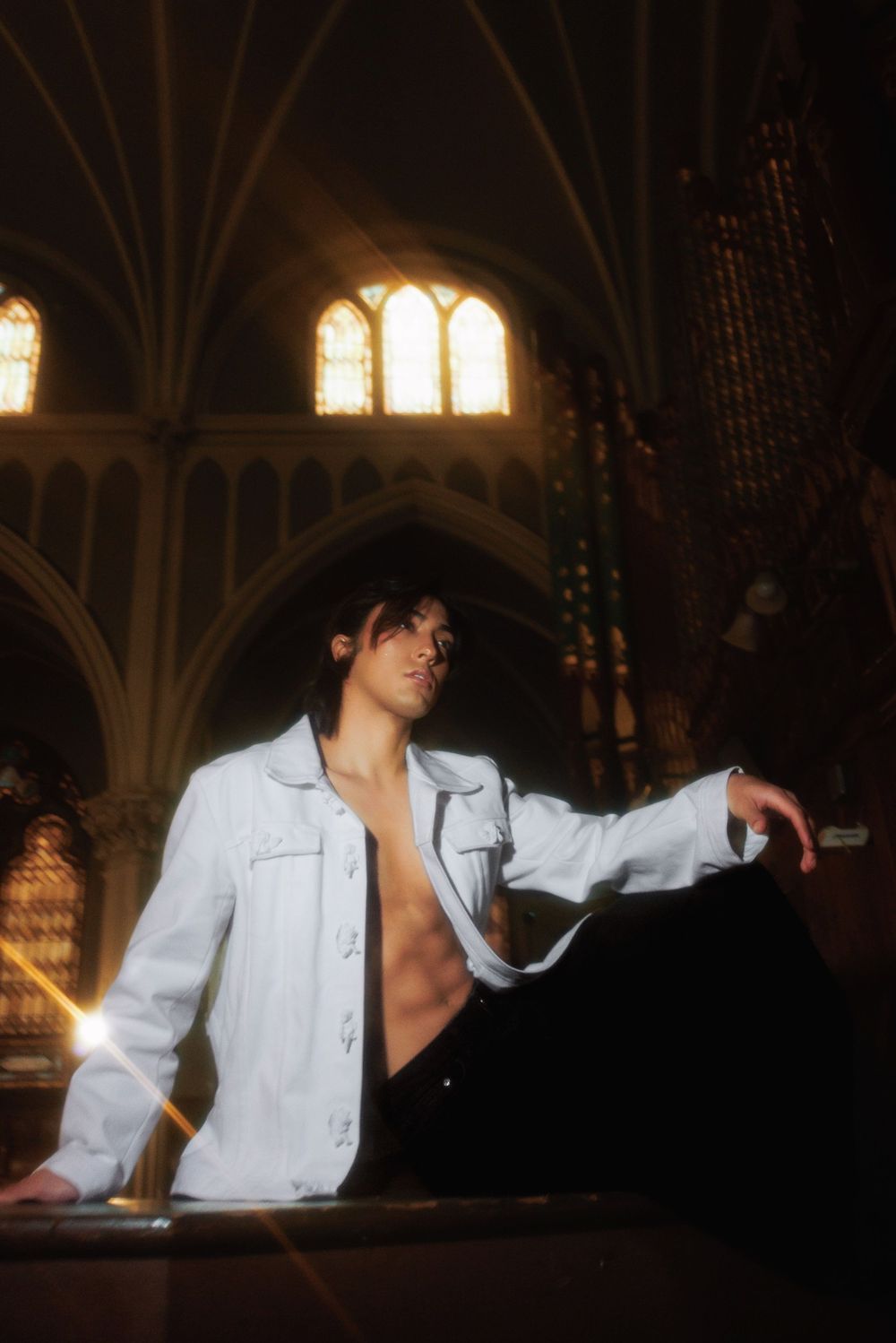 Figure in an open white shirt poses dramatically in a dimly lit church interior.