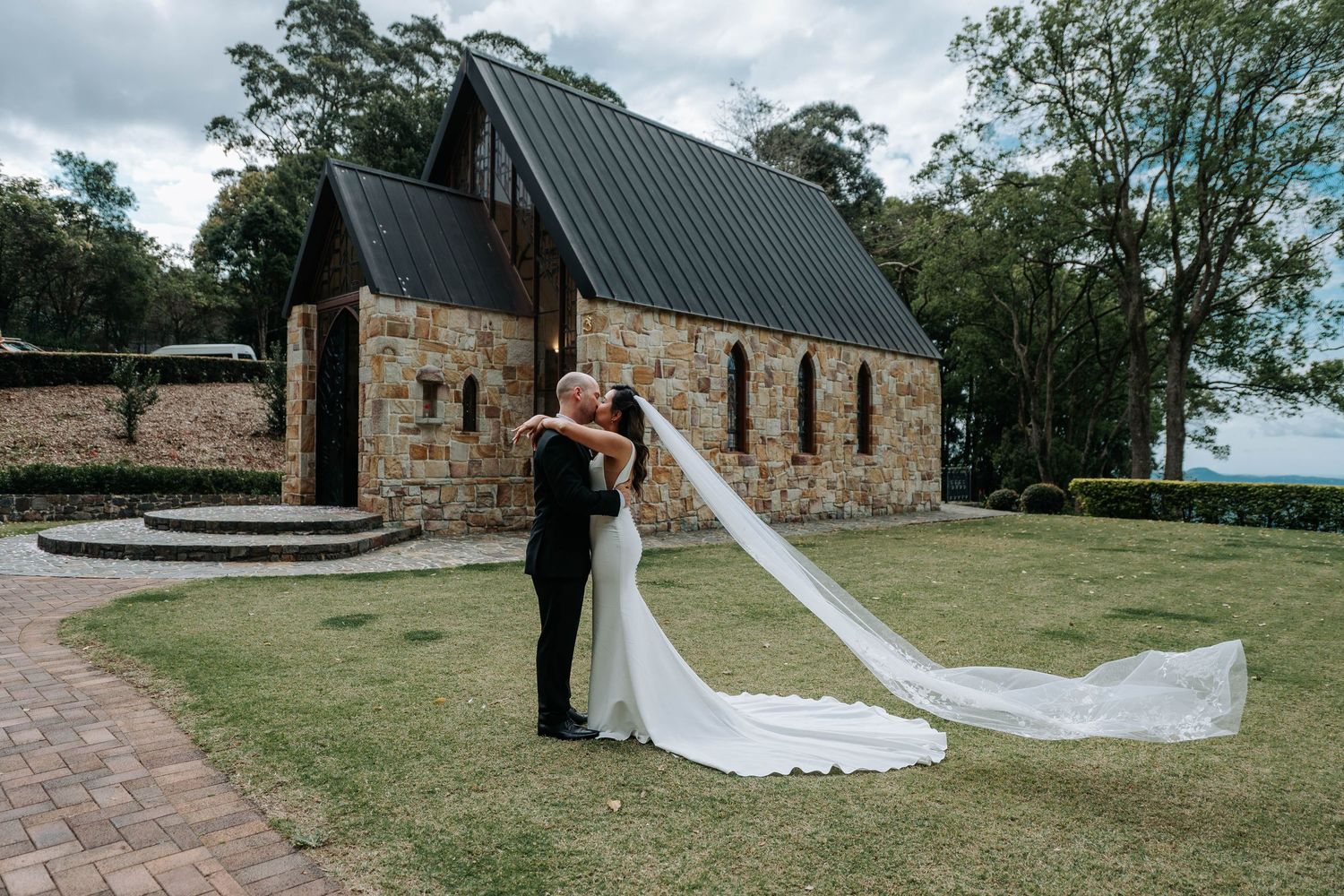 A couple shares a romantic moment outside a rustic stone chapel with a dramatic metal roof in a countryside setting.