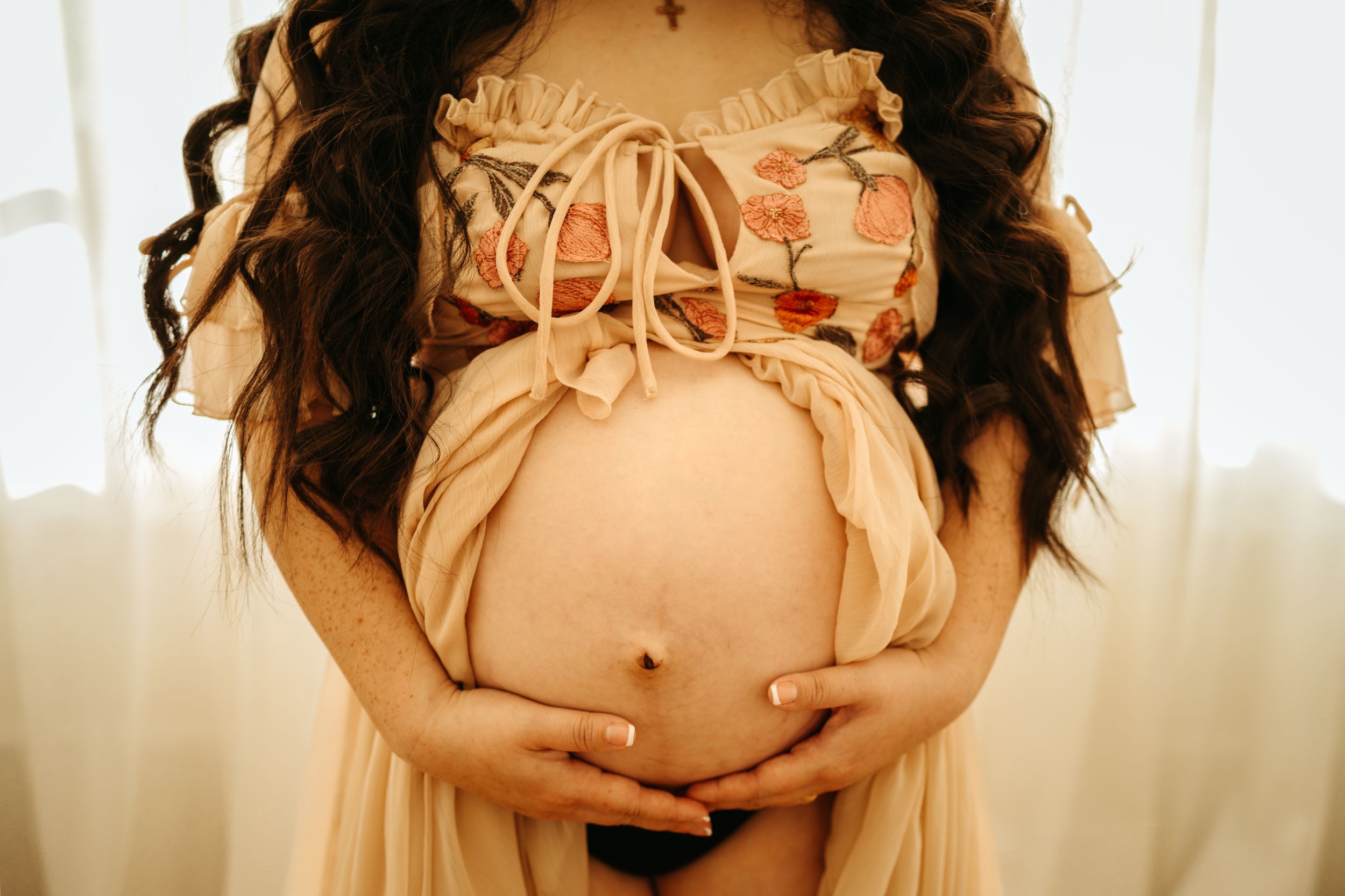 A pregnant woman in a peach-colored dress cradles her baby bump while standing in front of a window.