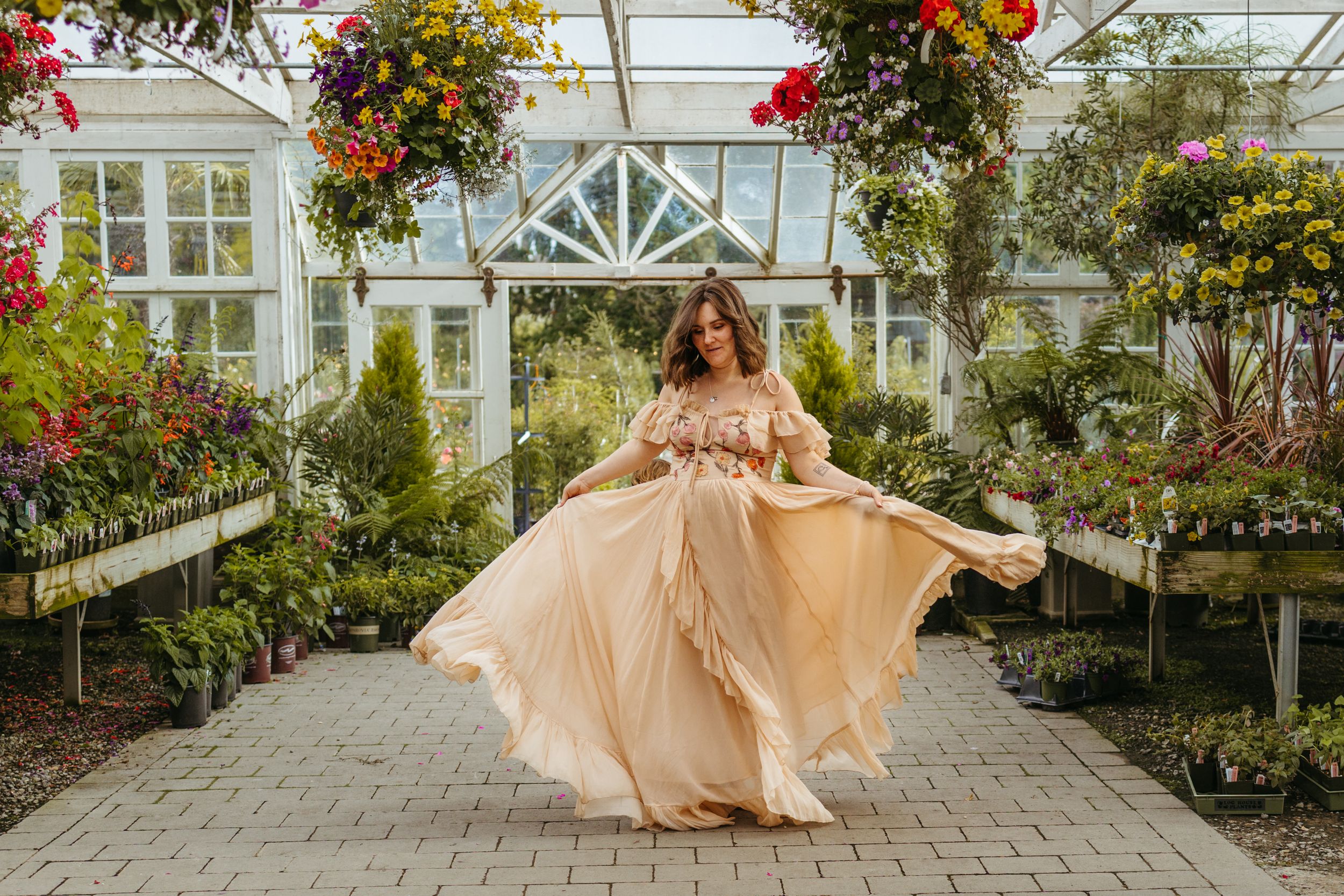 A person in a flowing peach gown poses in a greenhouse filled with vibrant hanging flowers and greenery.
