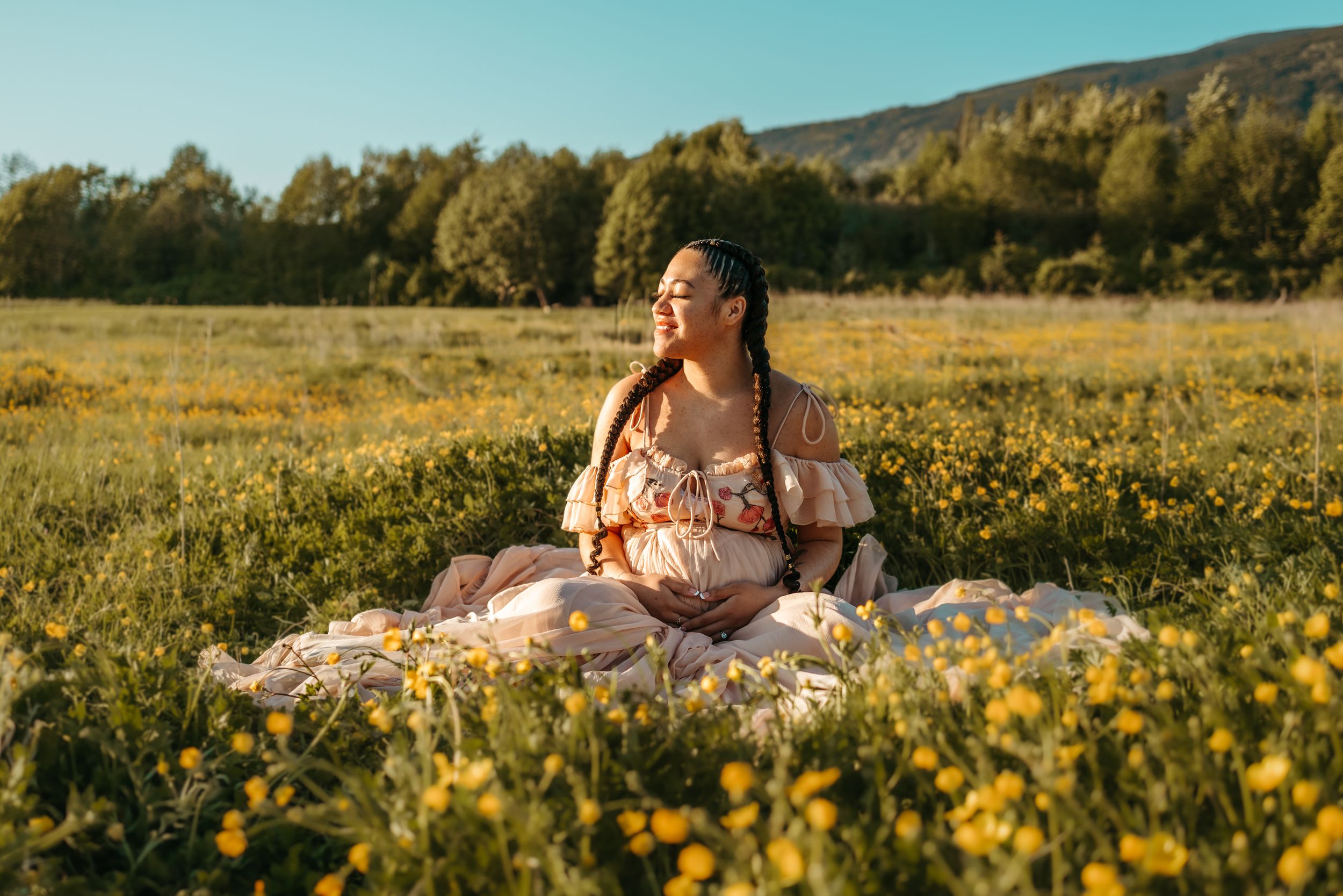 Someone in a floral dress lies in a golden wildflower meadow during sunset with mountains in the background.