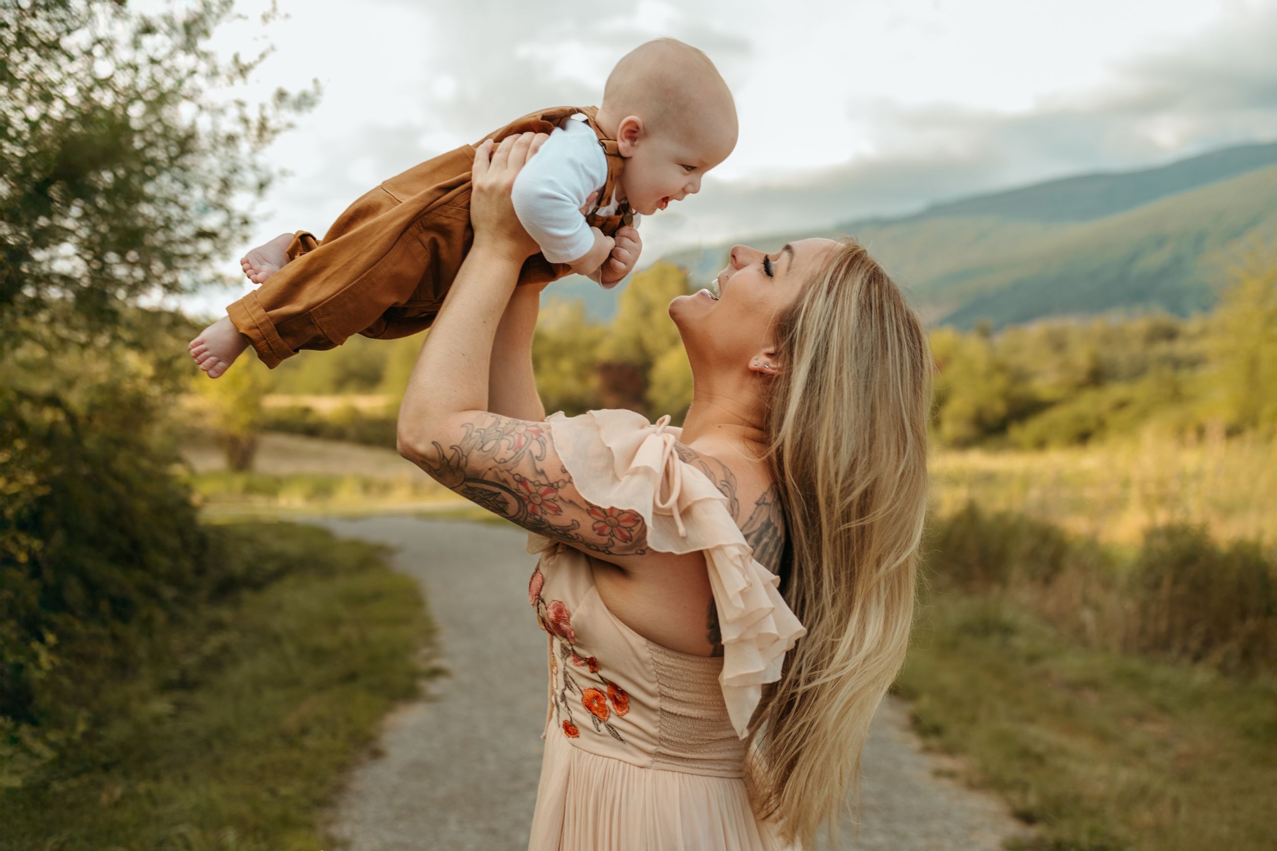 Parent in beige dress holds and snuggles with baby against scenic mountain landscape backdrop during sunset.