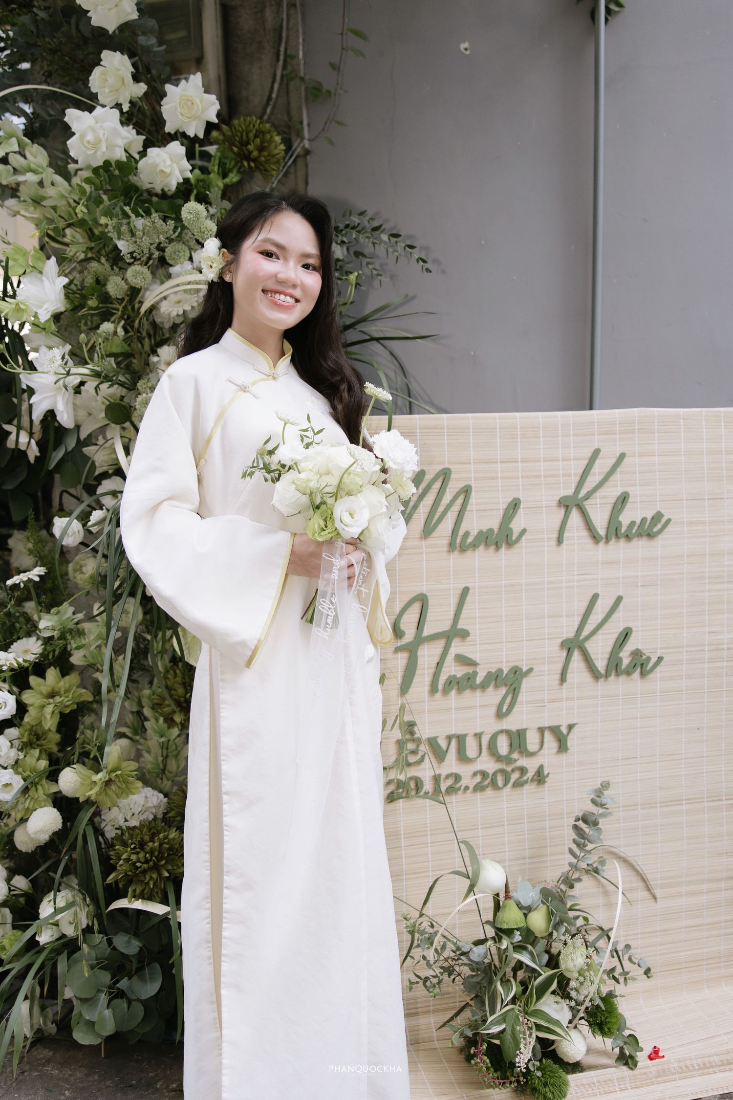 Smiling bride in white ao dai holding bouquet beside wedding sign reading Minh Khue Hoang Kha.