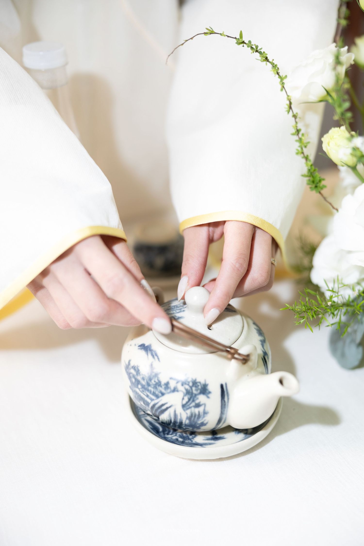 Close-up of bride in white ao dai pouring tea from blue and white porcelain teapot at wedding.