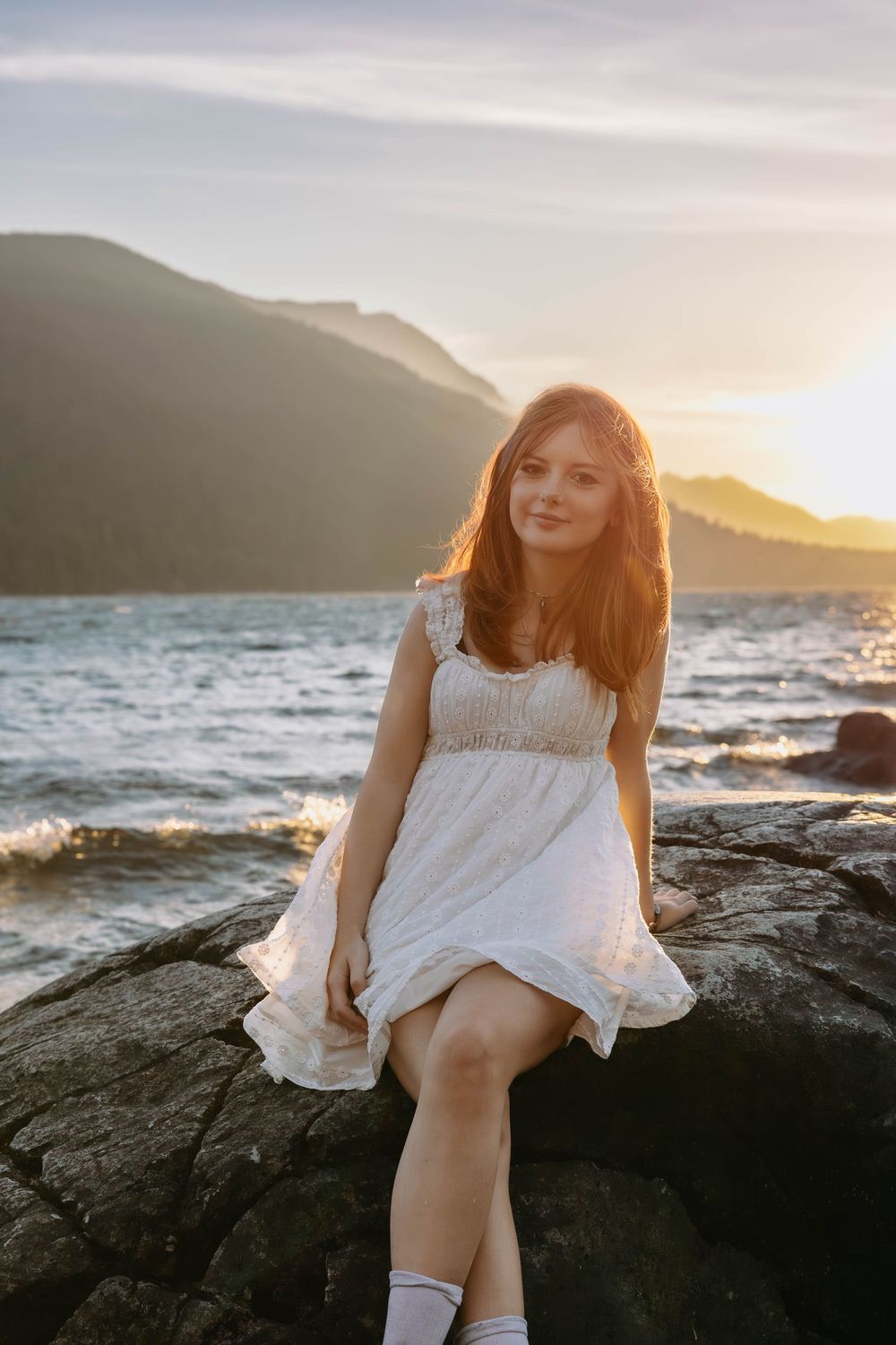 Person in white dress poses on coastal rocks at sunset with mountains in background.