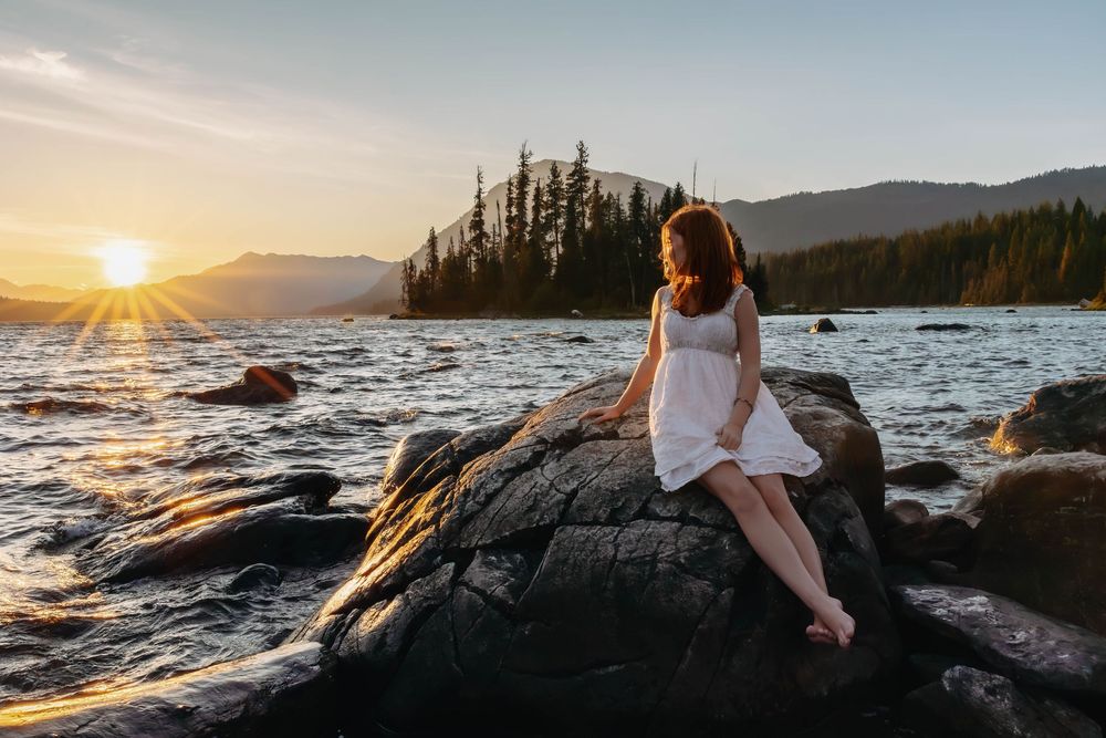 A dreamy sunset photo on rocky shoreline with mountains and evergreens in background.