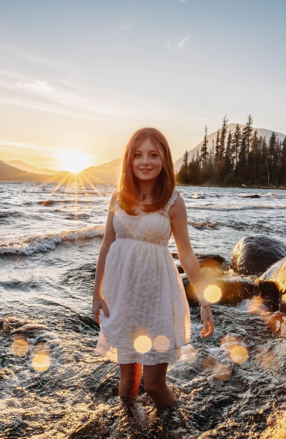 A white sundress-clad figure stands in shallow water against a mountain sunset with lens flares.