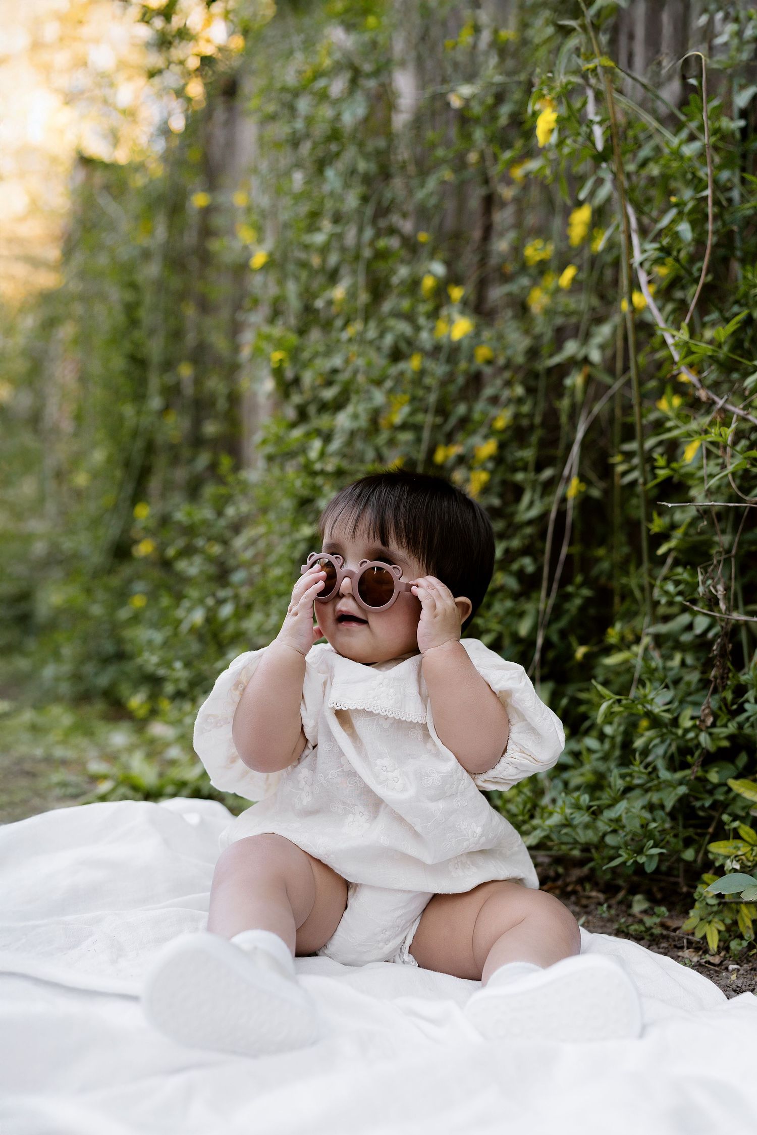 Small child wearing sunglasses and white dress sits on blanket outdoors surrounded by green foliage in natural light.