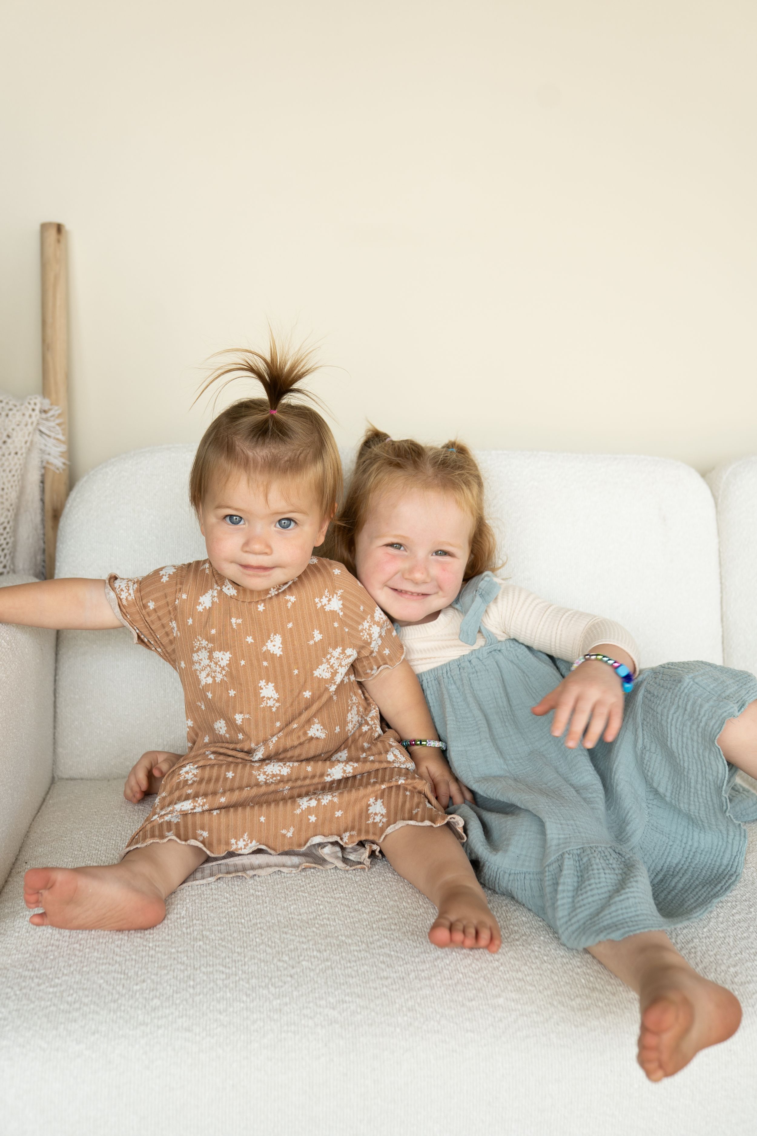 Two toddlers sit together on white couch wearing brown and blue dresses.