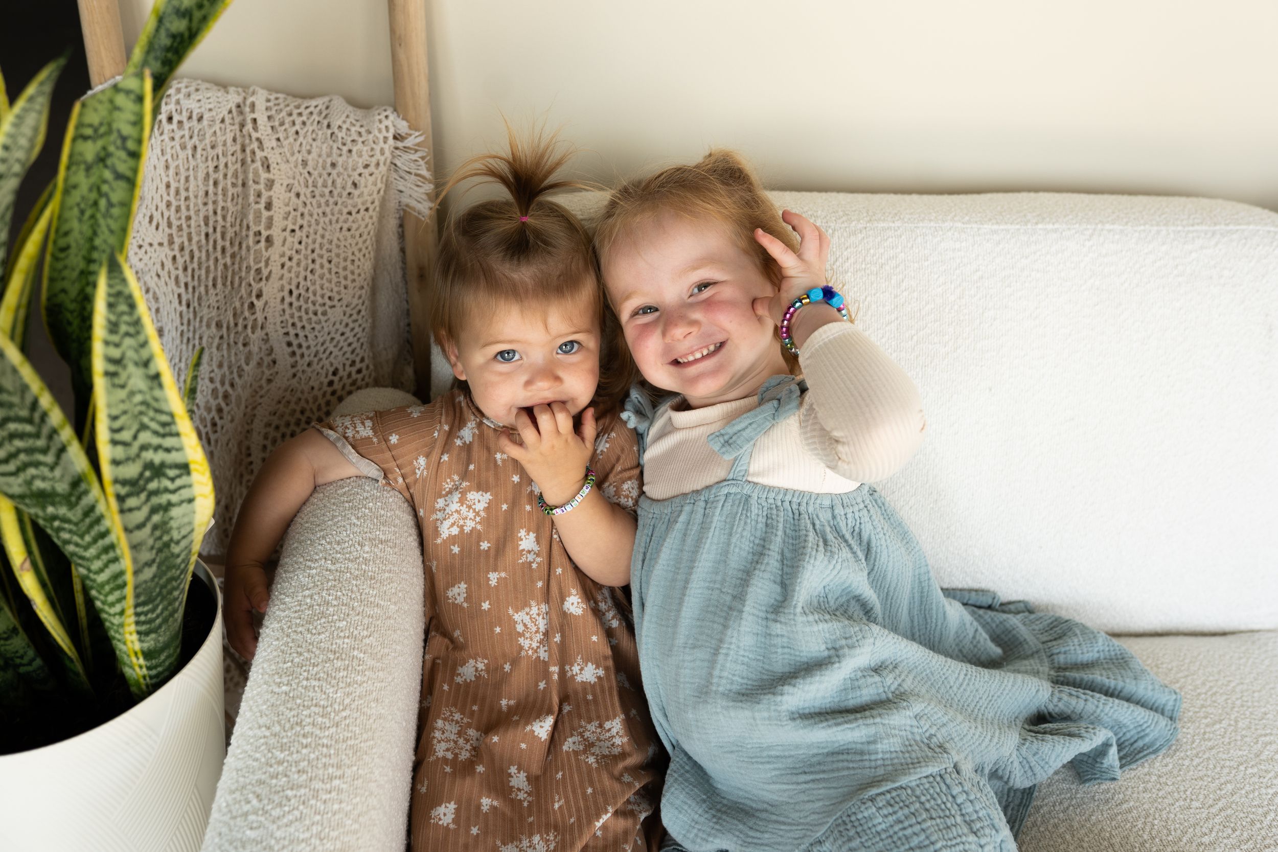 Two children sitting together on a white couch with decorative pillows and a plant in the background.