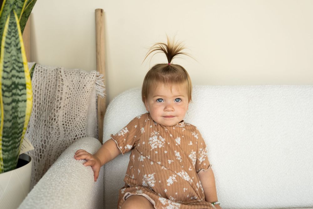 A toddler with a hair bow wearing a brown dress sitting on a couch.