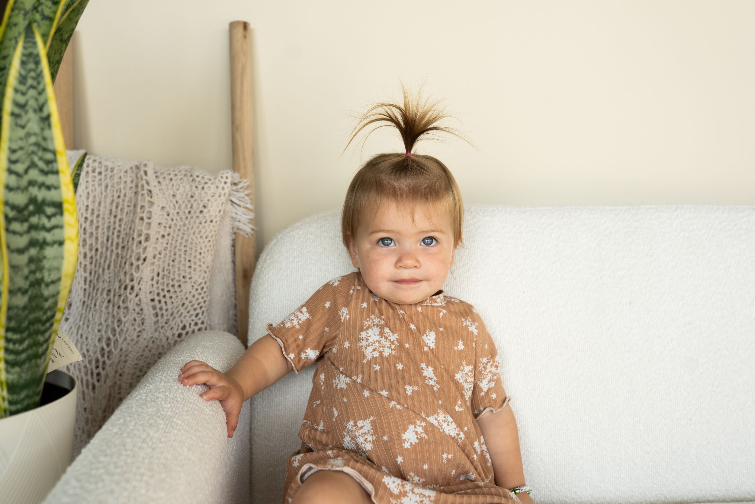 A toddler with a hair bow wearing a brown dress sitting on a couch.