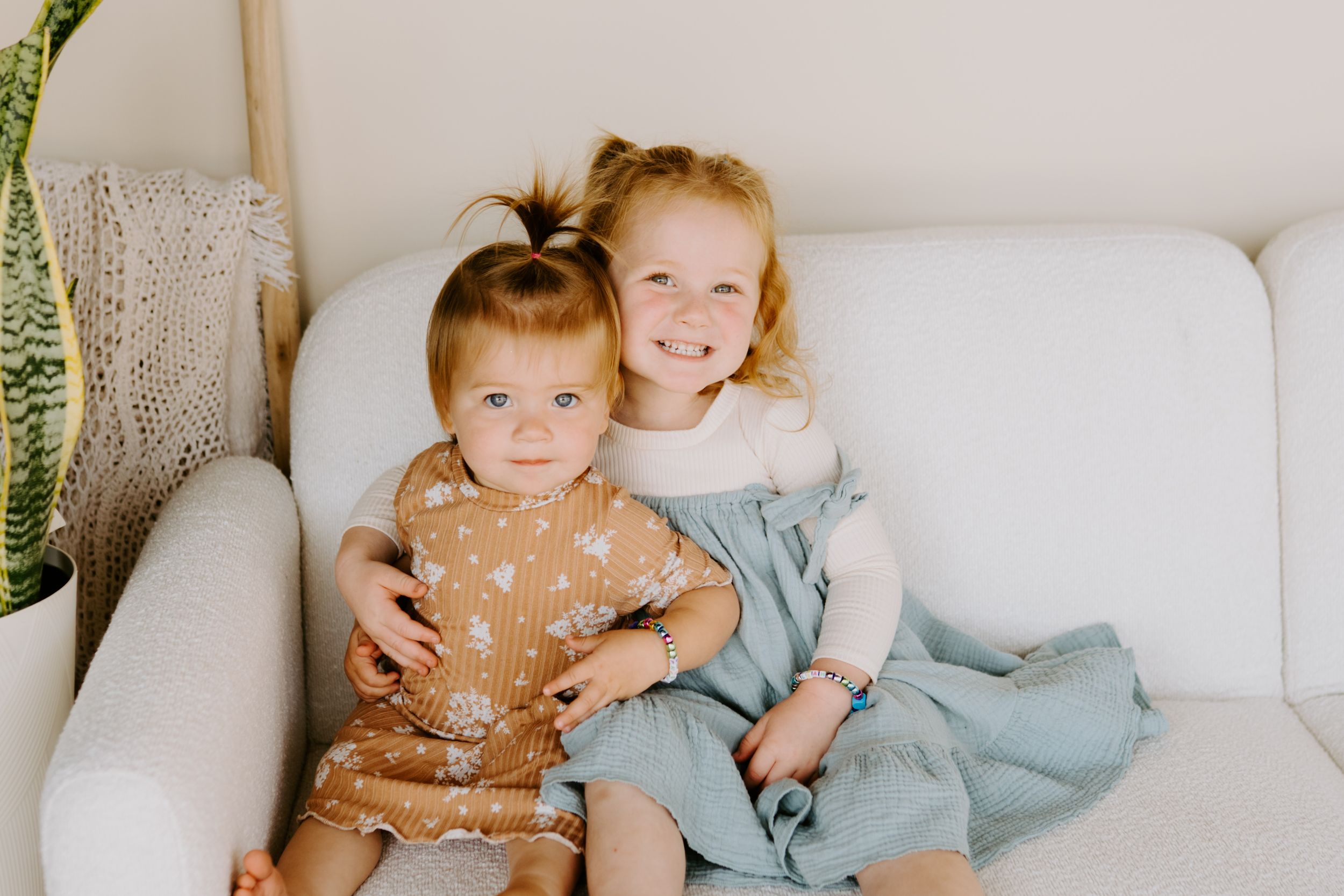 Two young children sitting together on a white couch with a plant visible.