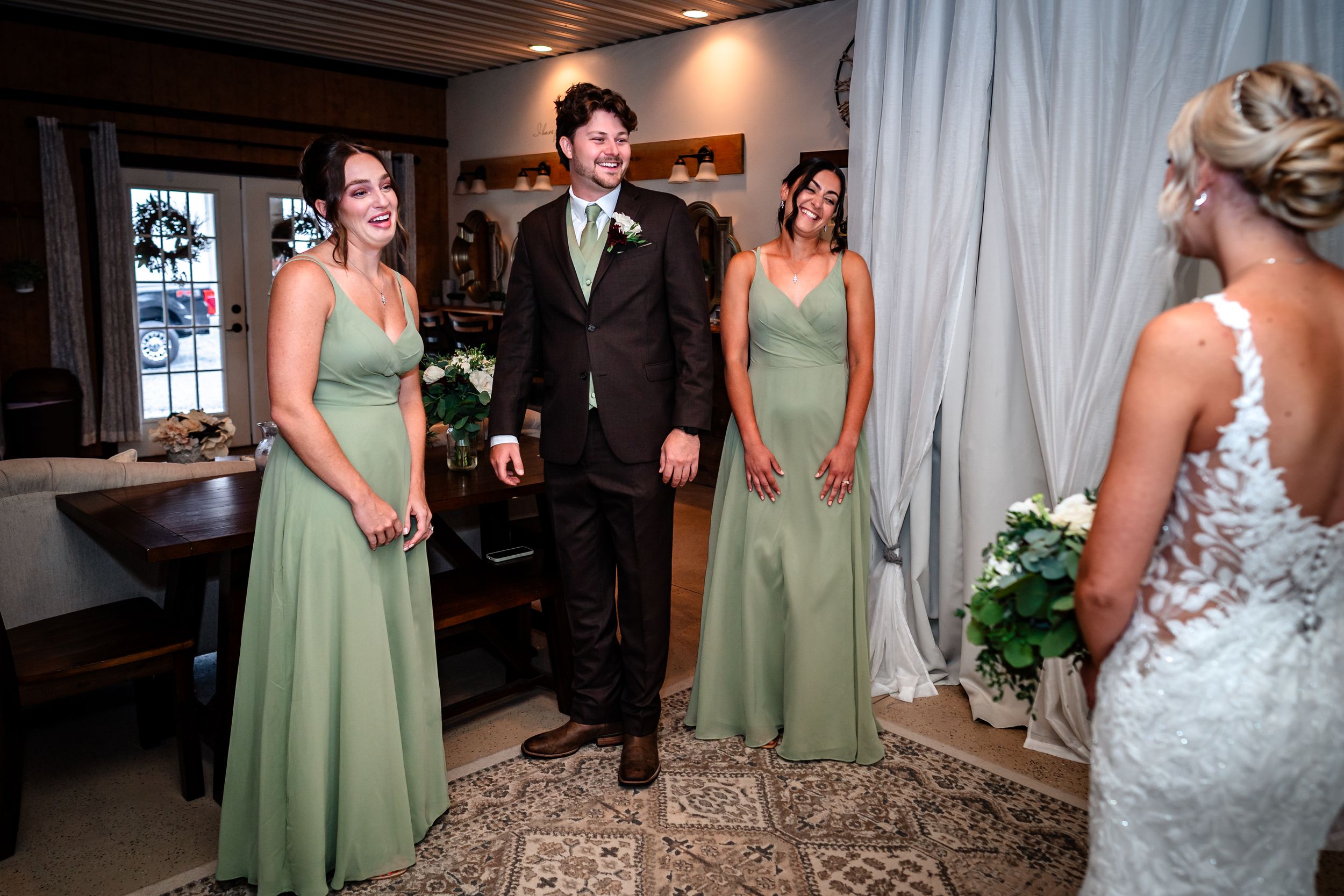 Bridesmaids in sage green dresses walk down a hallway with white walls and dark wood floors.