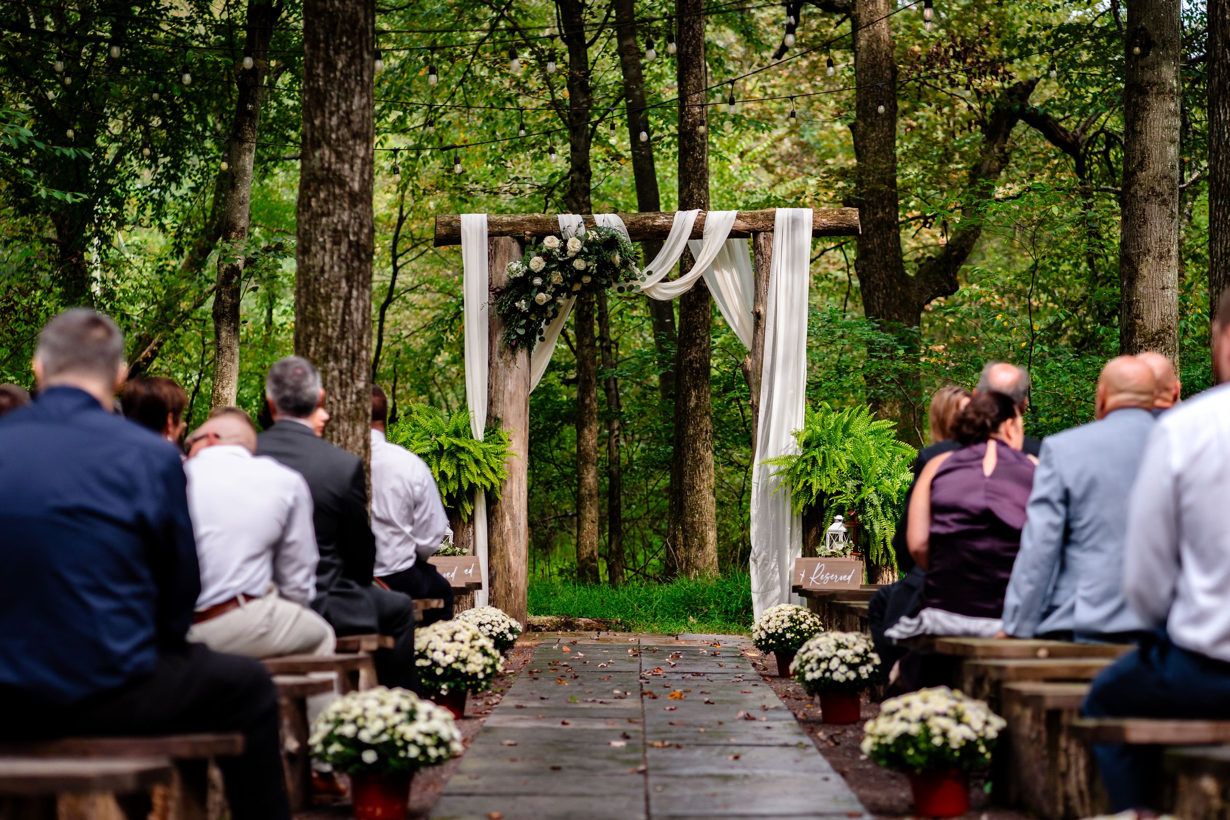 Wedding ceremony in a lush forest setting with white floral arrangements and wooden benches.