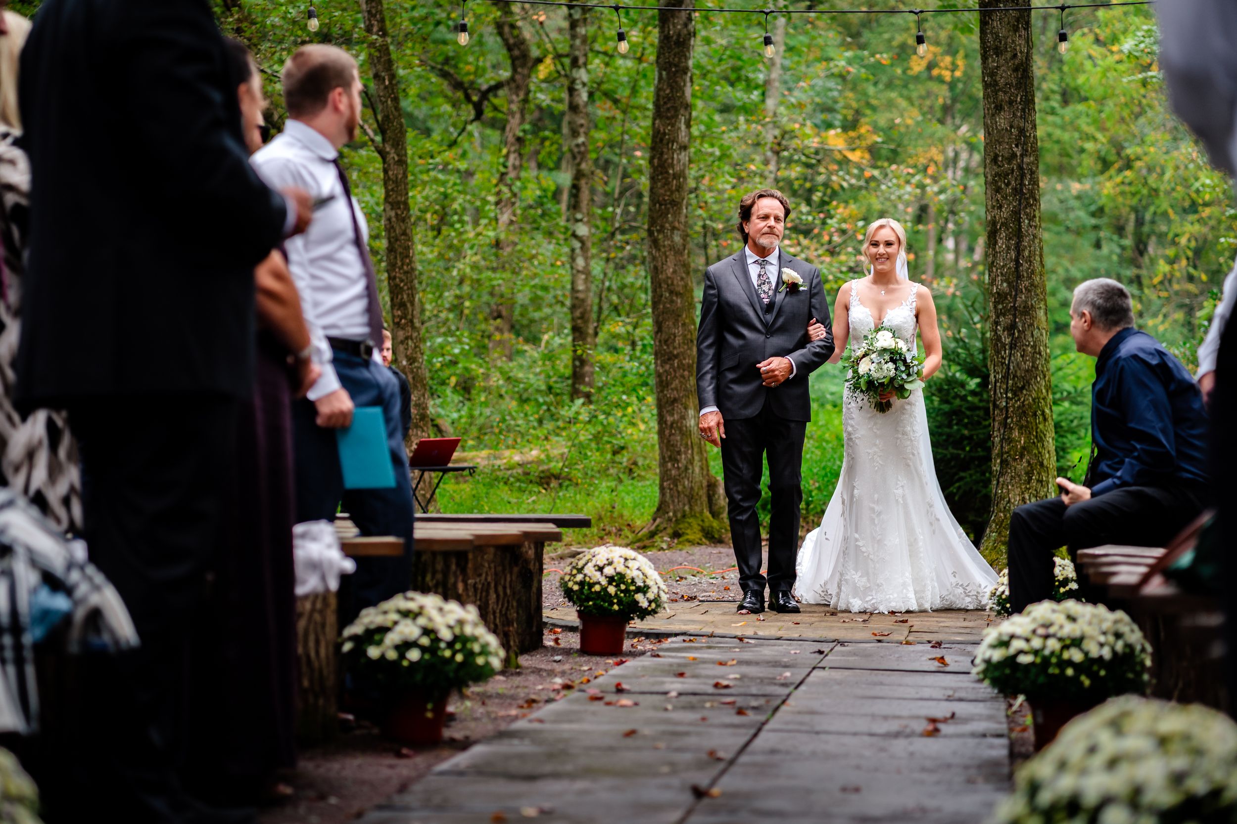 Outdoor wedding ceremony in progress along a wooden pathway surrounded by green forest.