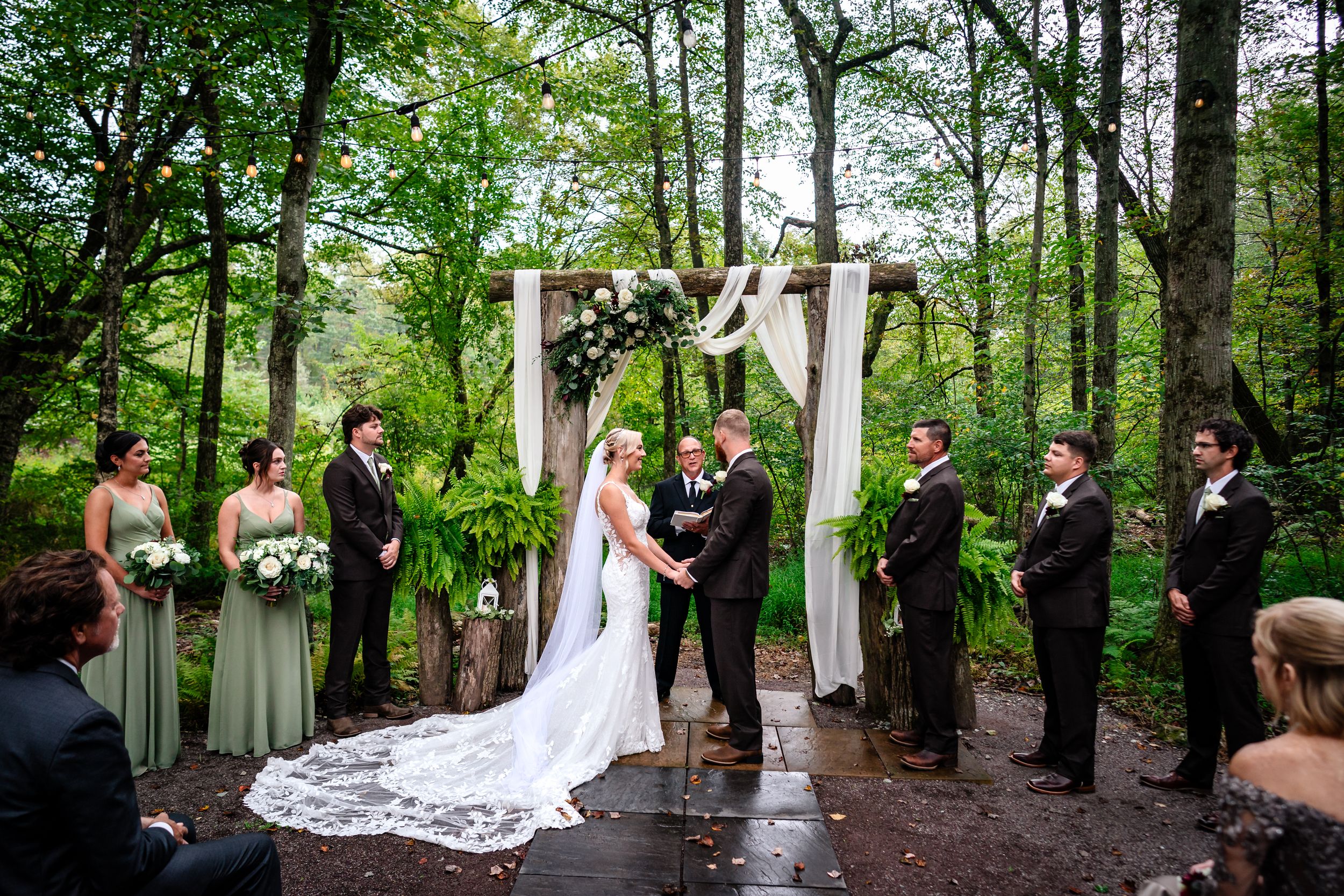 Wedding ceremony taking place under a decorated wooden arch in a lush forest setting with flowing white fabric and florals.