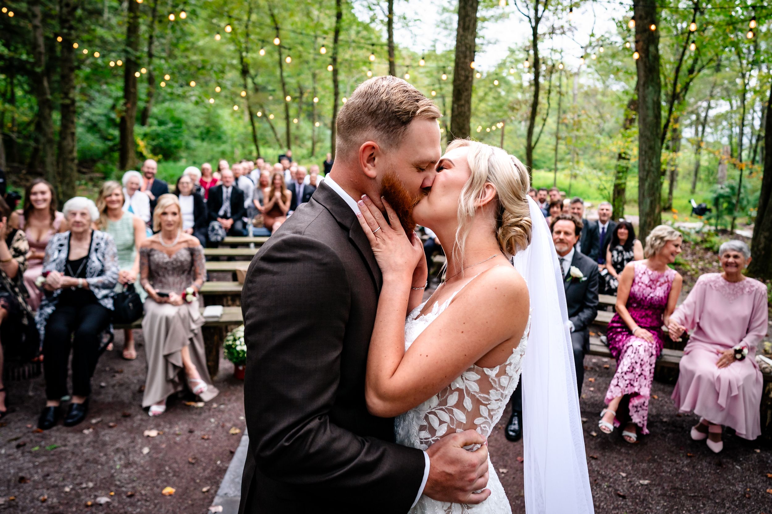 Intimate wedding kiss moment surrounded by seated guests in a wooded outdoor ceremony space.