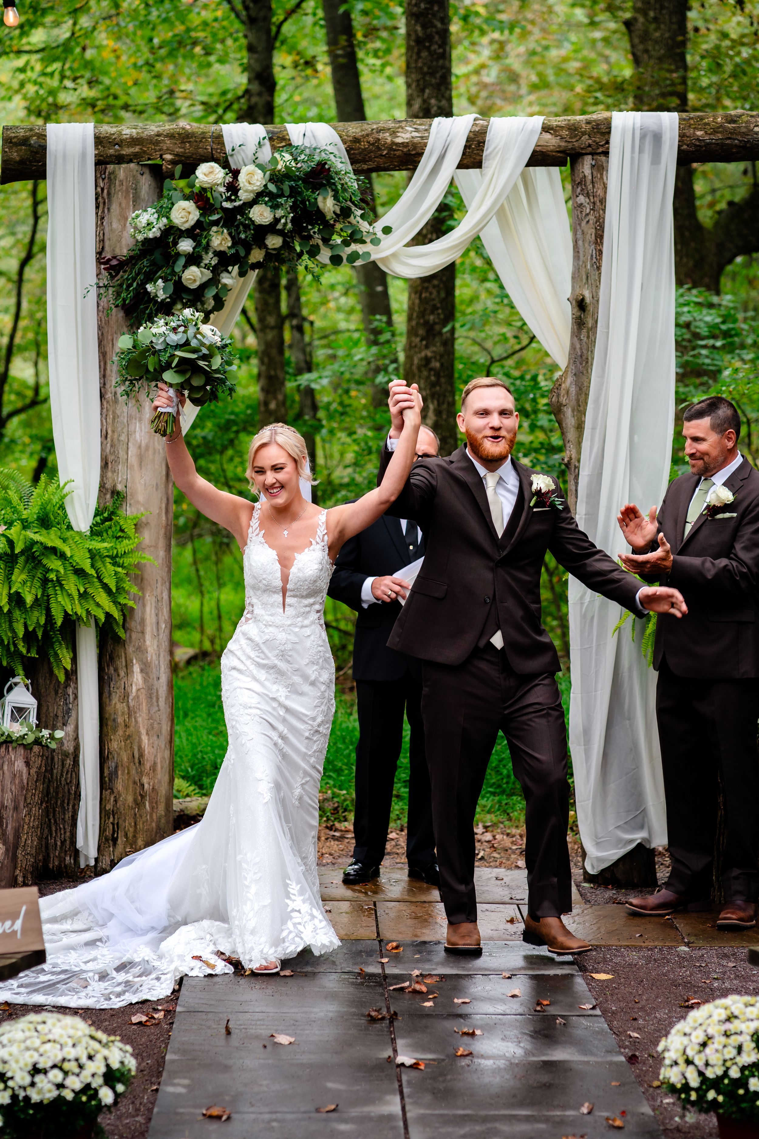 Celebratory moment under rustic wooden arch decorated with white fabric and florals during outdoor wedding ceremony.