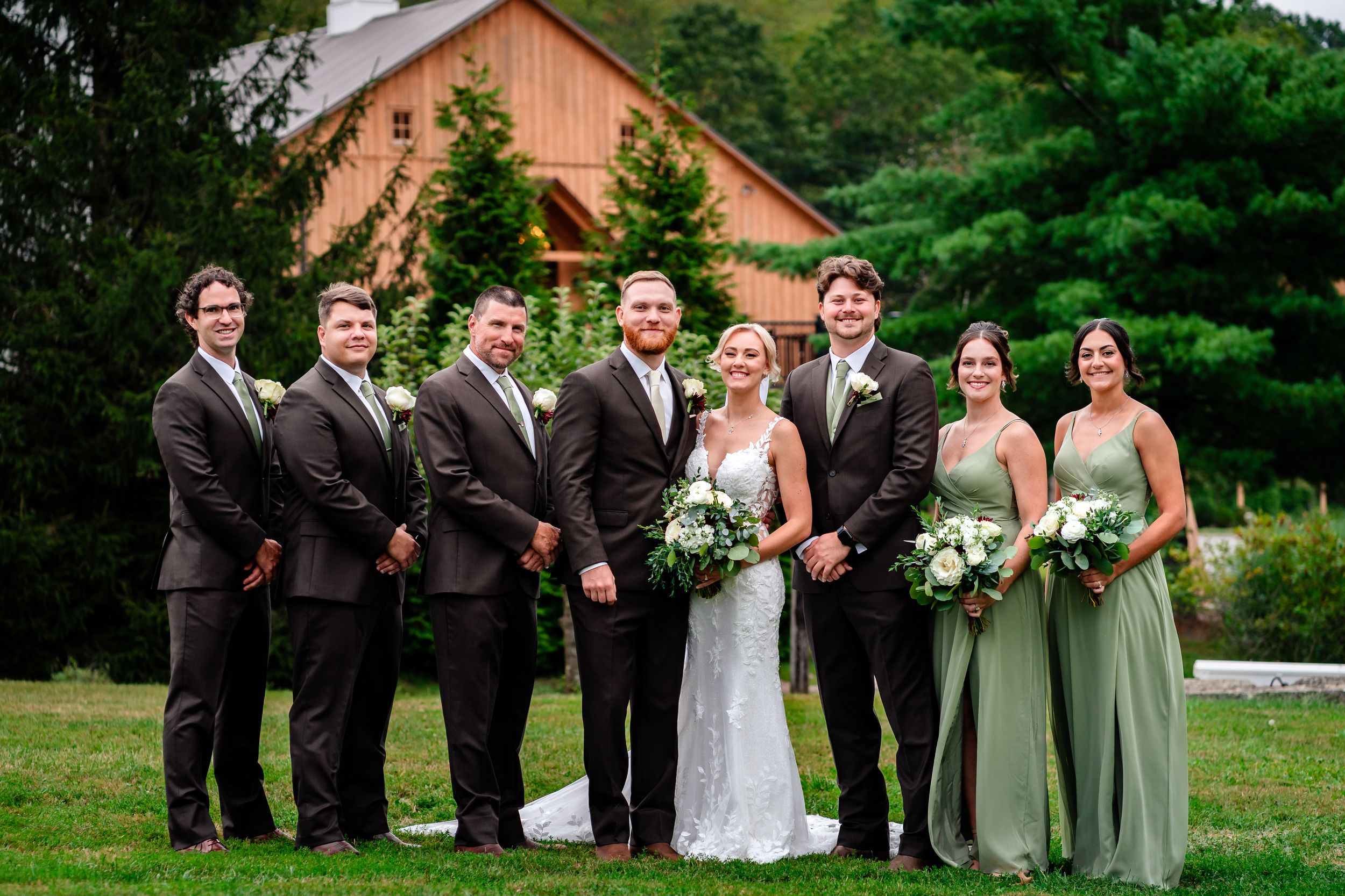 Wedding party in formal attire poses together in front of rustic barn with green mountain backdrop.