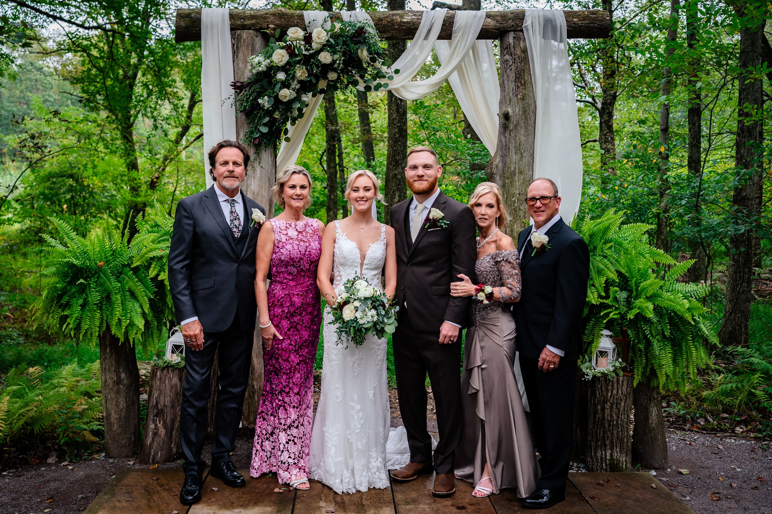 Wedding party poses under decorated wooden arch with ferns and white floral arrangements in forest setting.