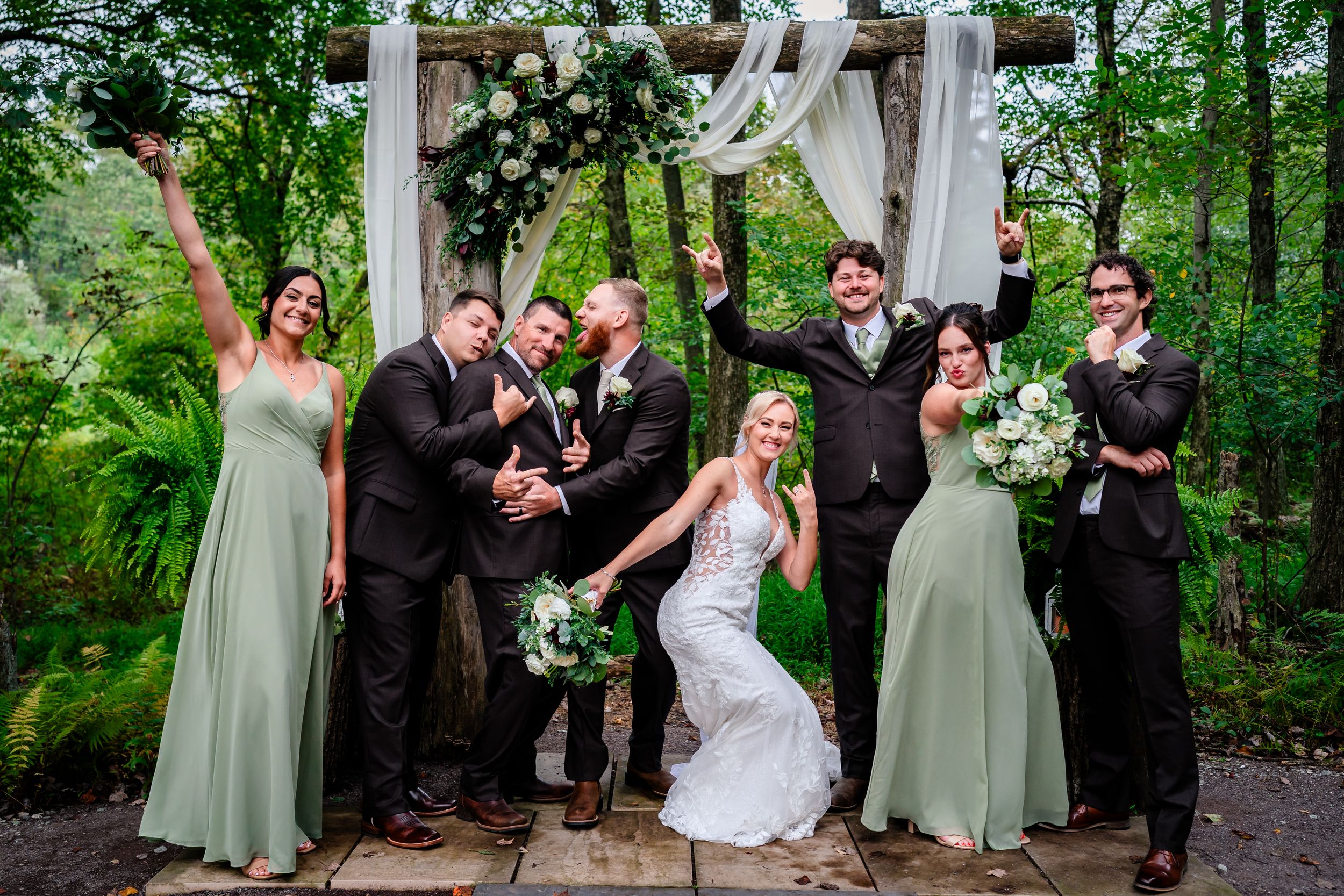 Fun wedding party photo with playful poses under decorated wooden arch in forest setting.