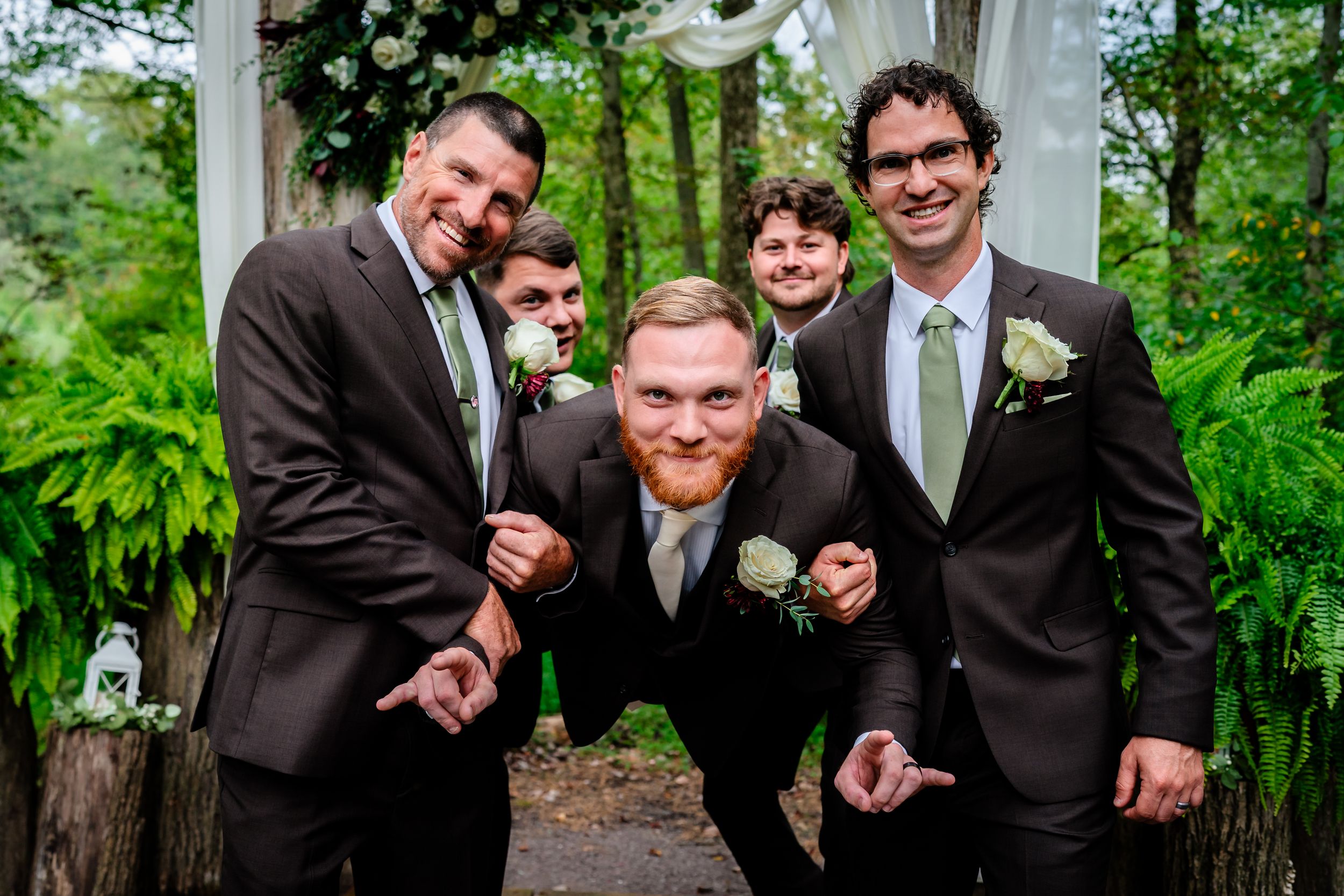 Groomsmen in dark suits pose playfully for photo on outdoor stone patio.