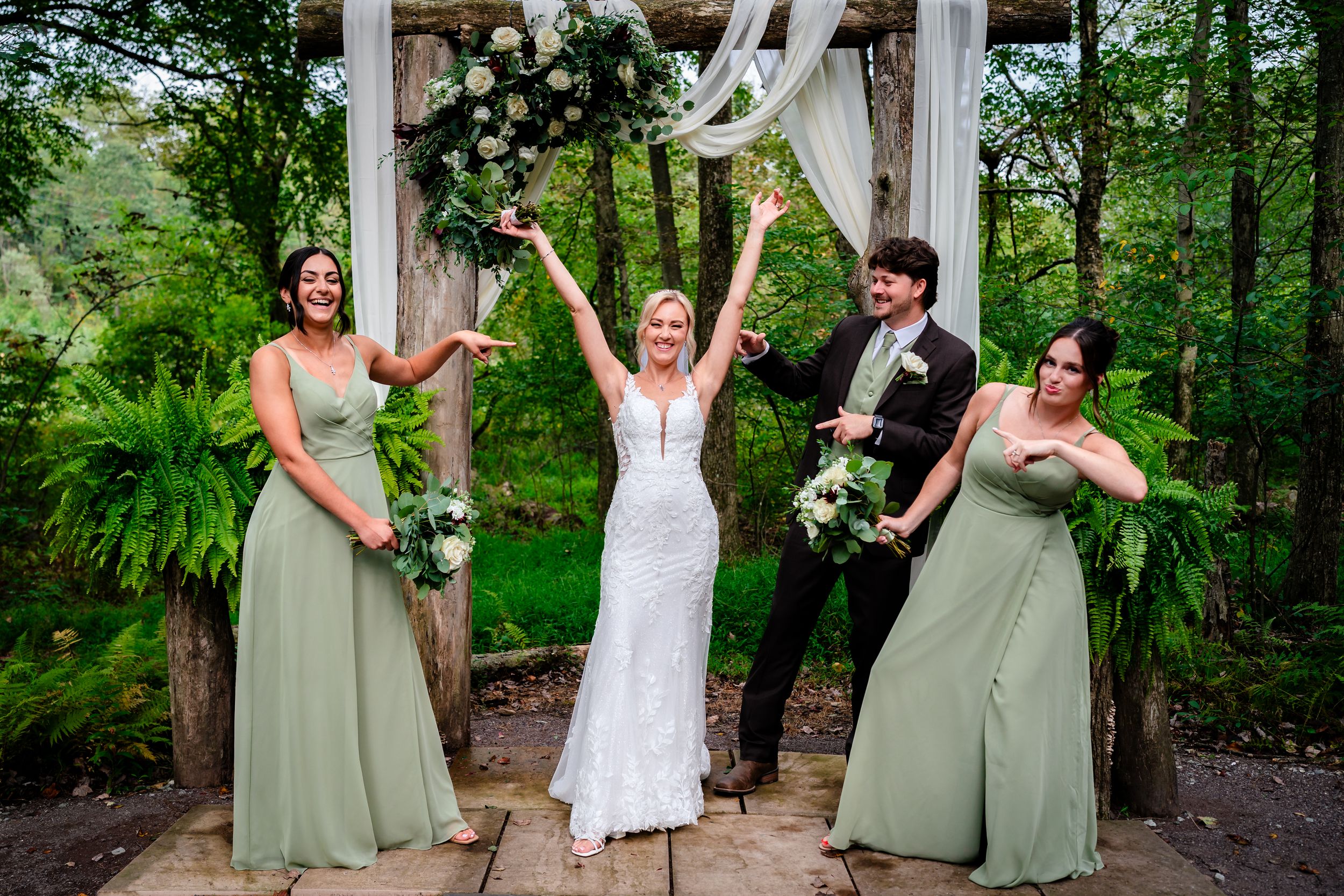Wedding party in sage green dresses and dark suits celebrating joyfully under decorated wooden arch.