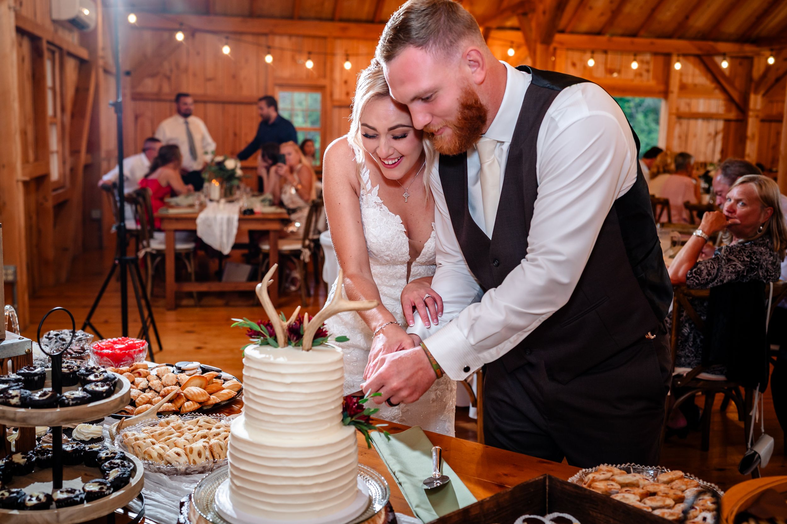 Wedding cake cutting ceremony in rustic barn venue with string lights overhead.