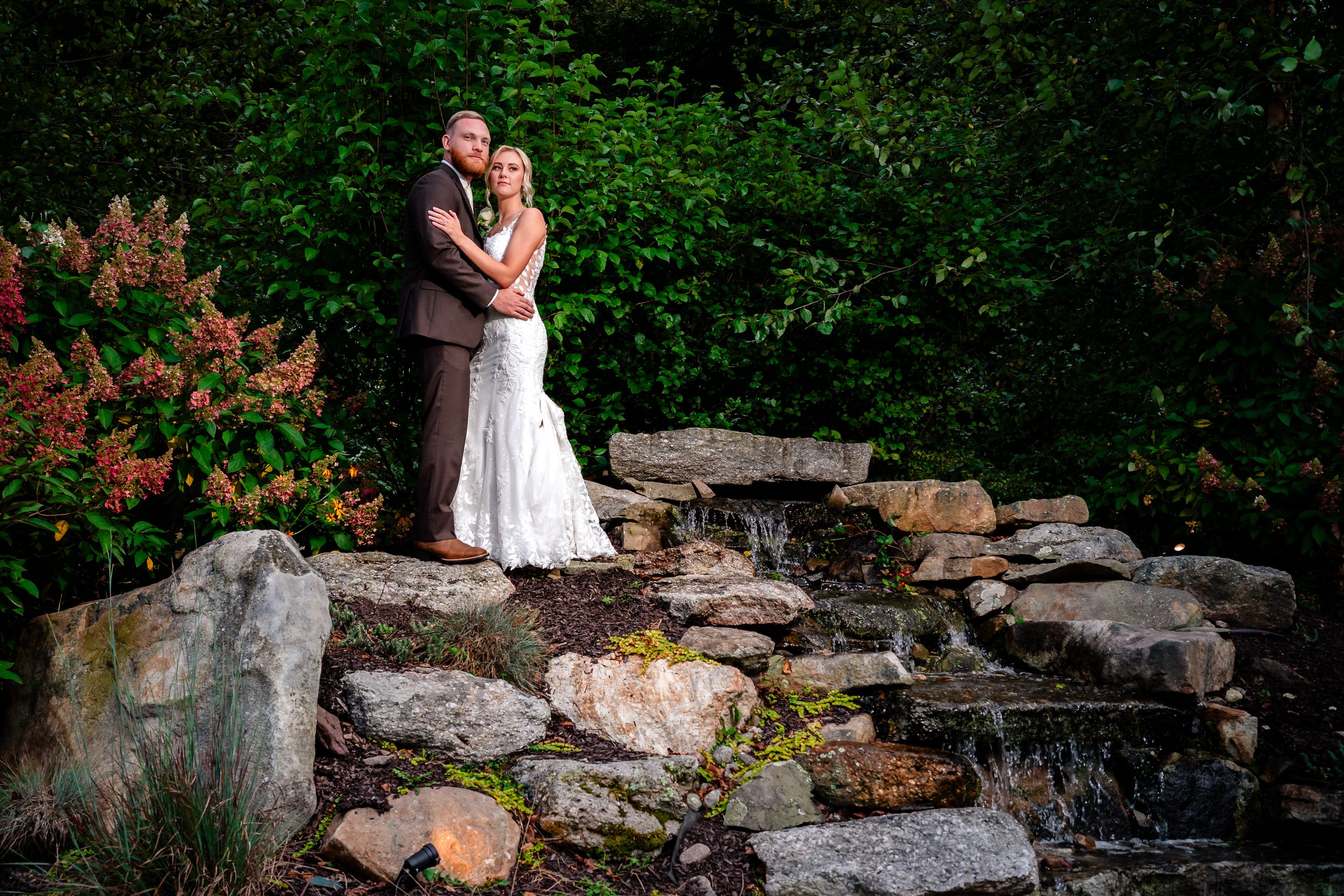Newlyweds posing on stone wall surrounded by lush summer greenery.