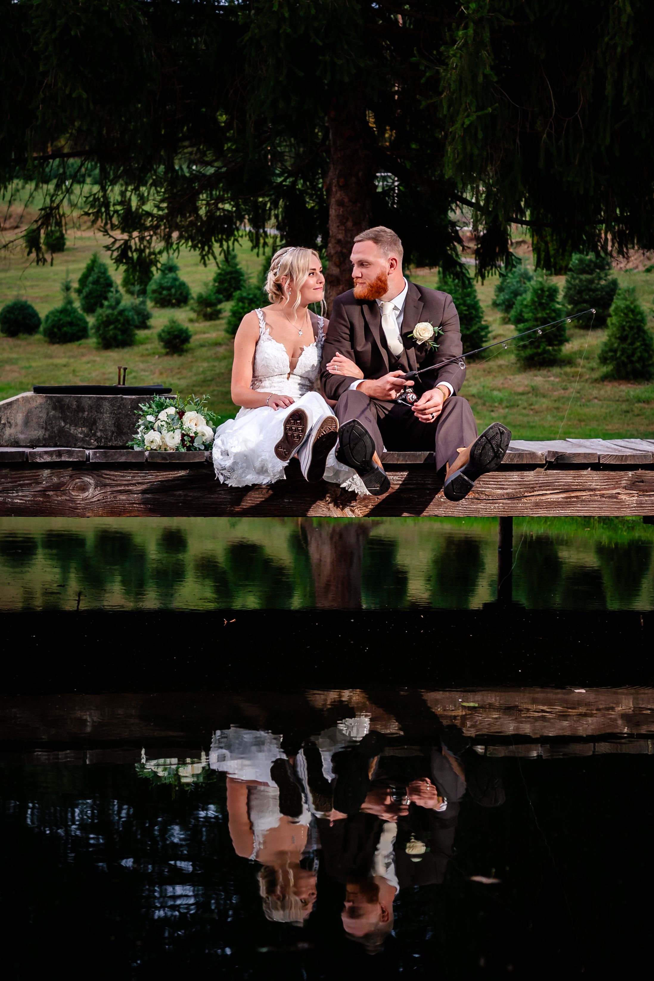 Wedding couple sitting by reflective pond creating mirror image in natural setting.
