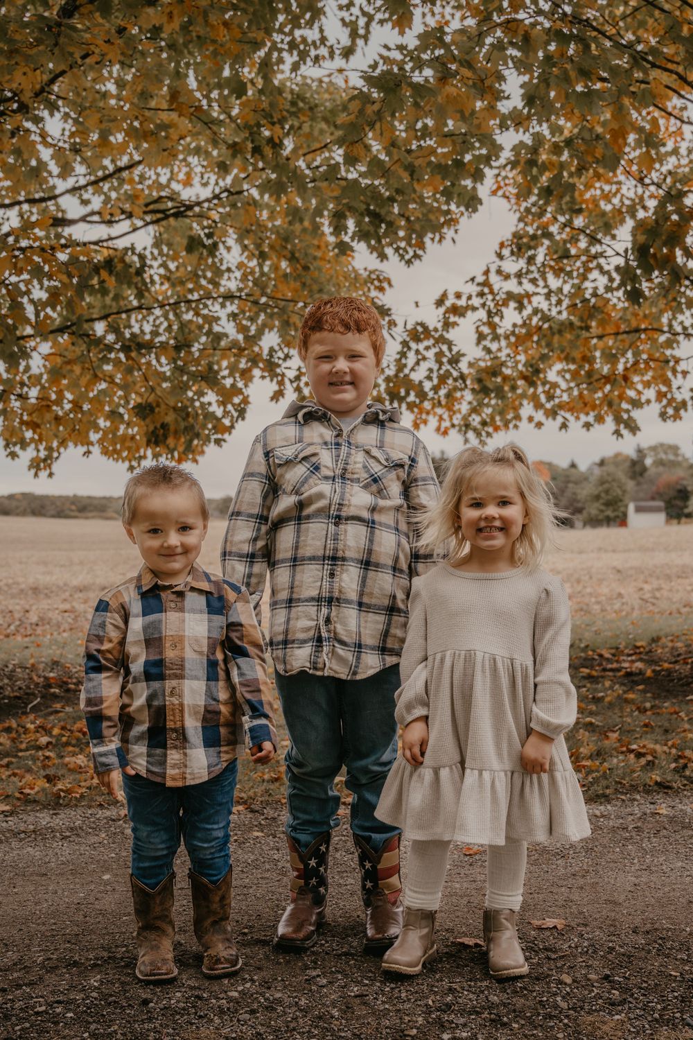 Three young children in fall plaid outfits stand together under autumn foliage on a rural path.