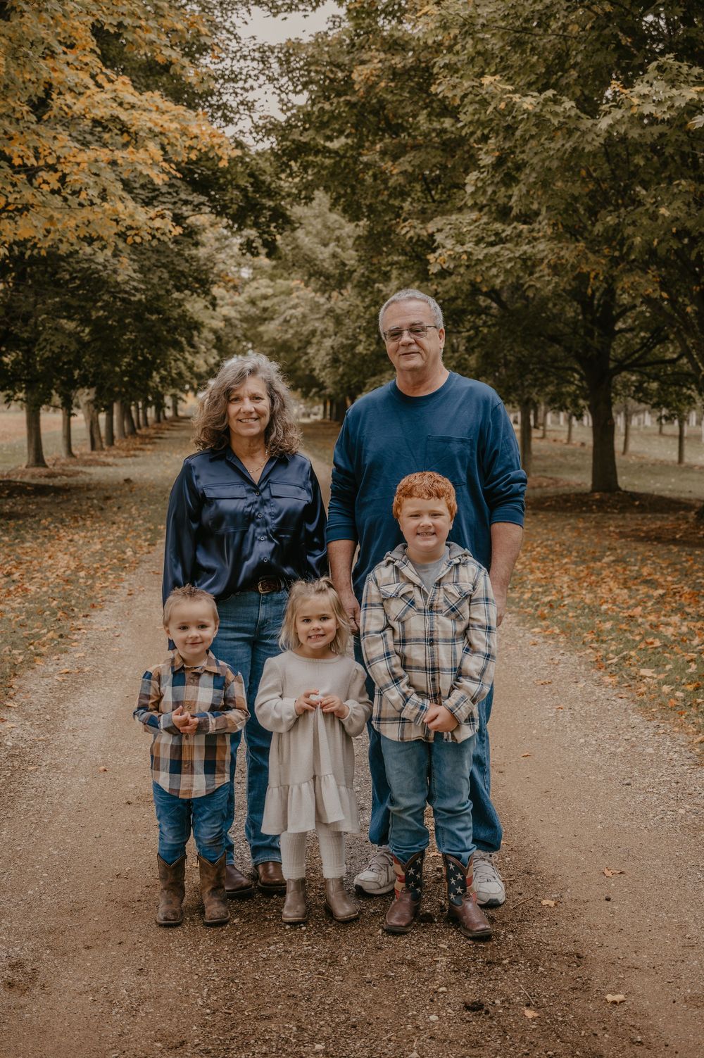 Grandparents pose with three young grandchildren on a scenic tree-lined path in fall foliage.