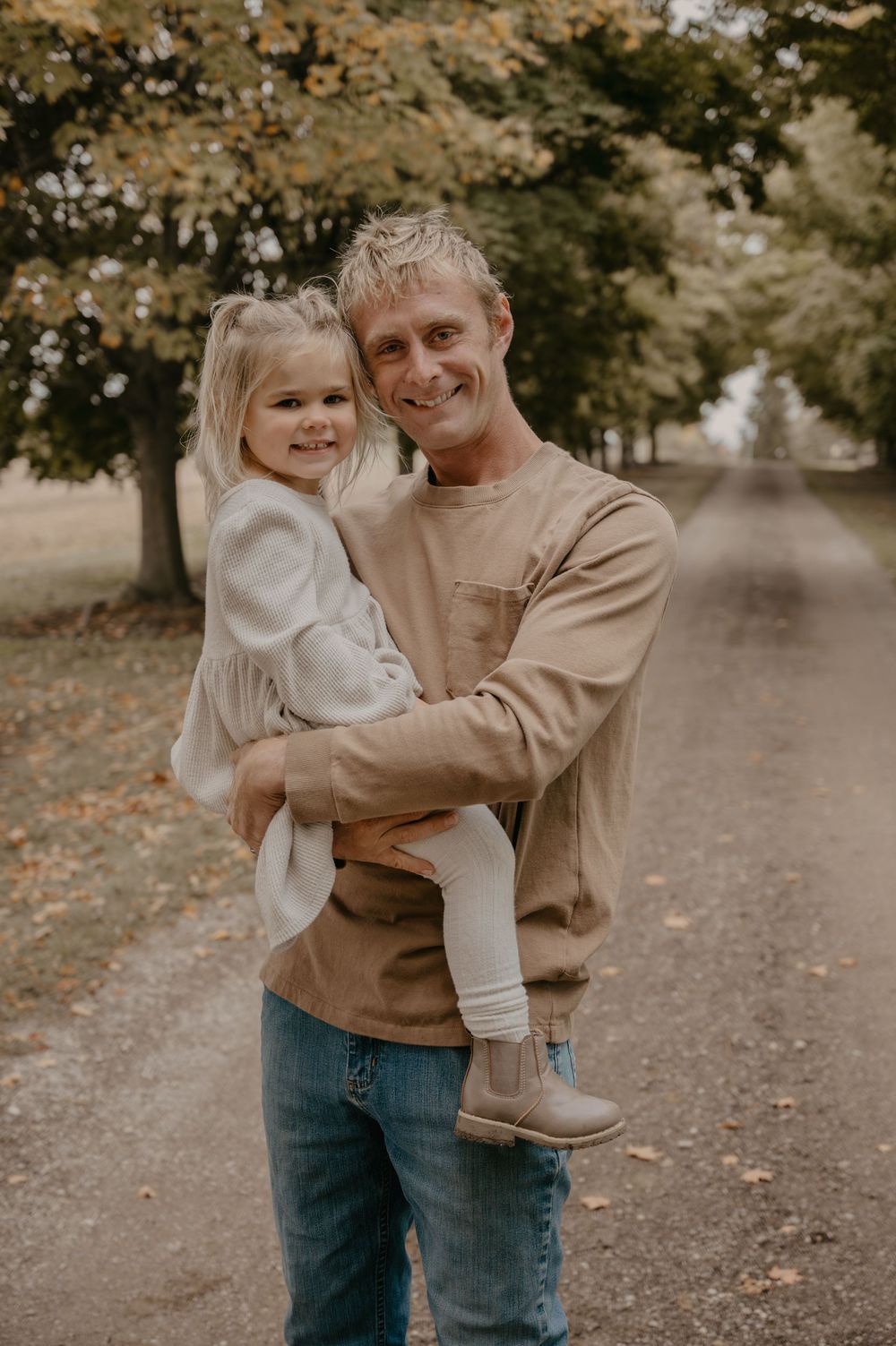 Smiling father holds young blonde daughter on a scenic autumn tree-lined country road.