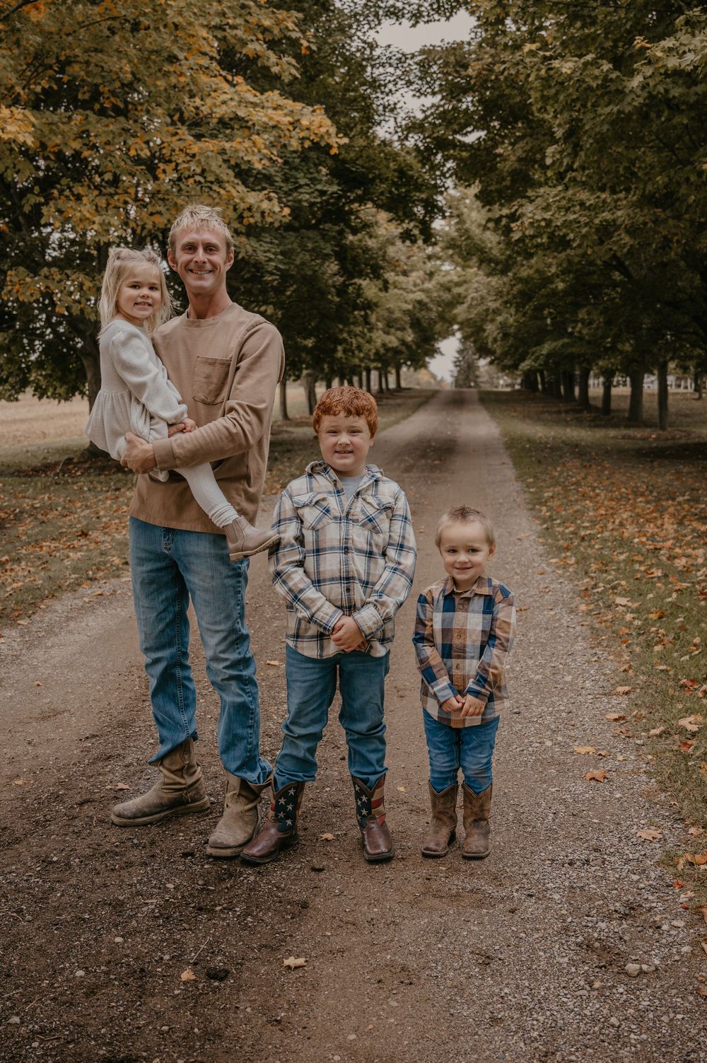 Father holds daughter while posing with two sons on a tree-lined path during fall family photos.