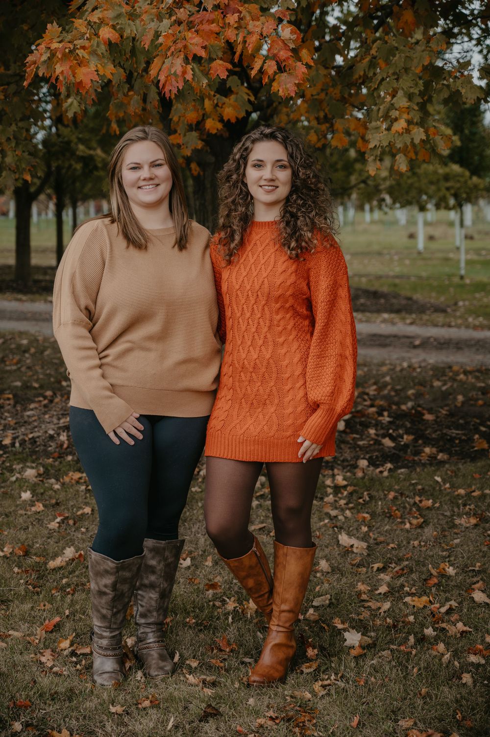 Two young women smile together wearing camel and orange sweaters beneath vibrant autumn foliage.