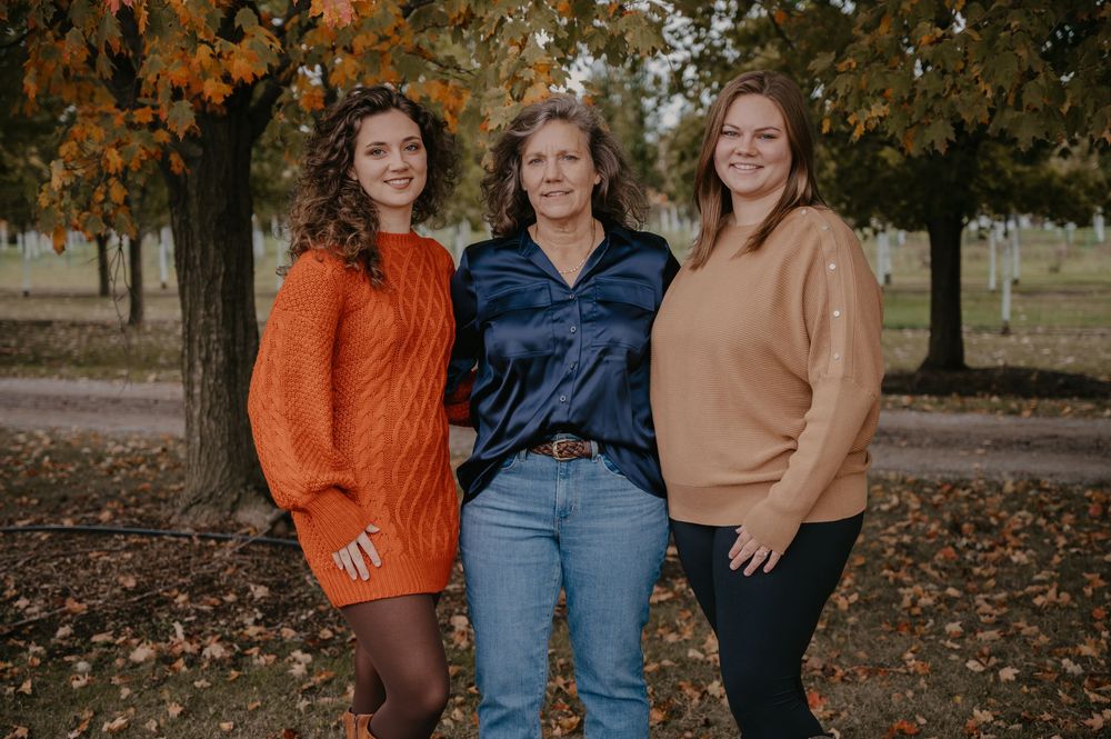 Three women pose together in an autumn park, wearing orange, navy, and tan outfits among colorful fall foliage.