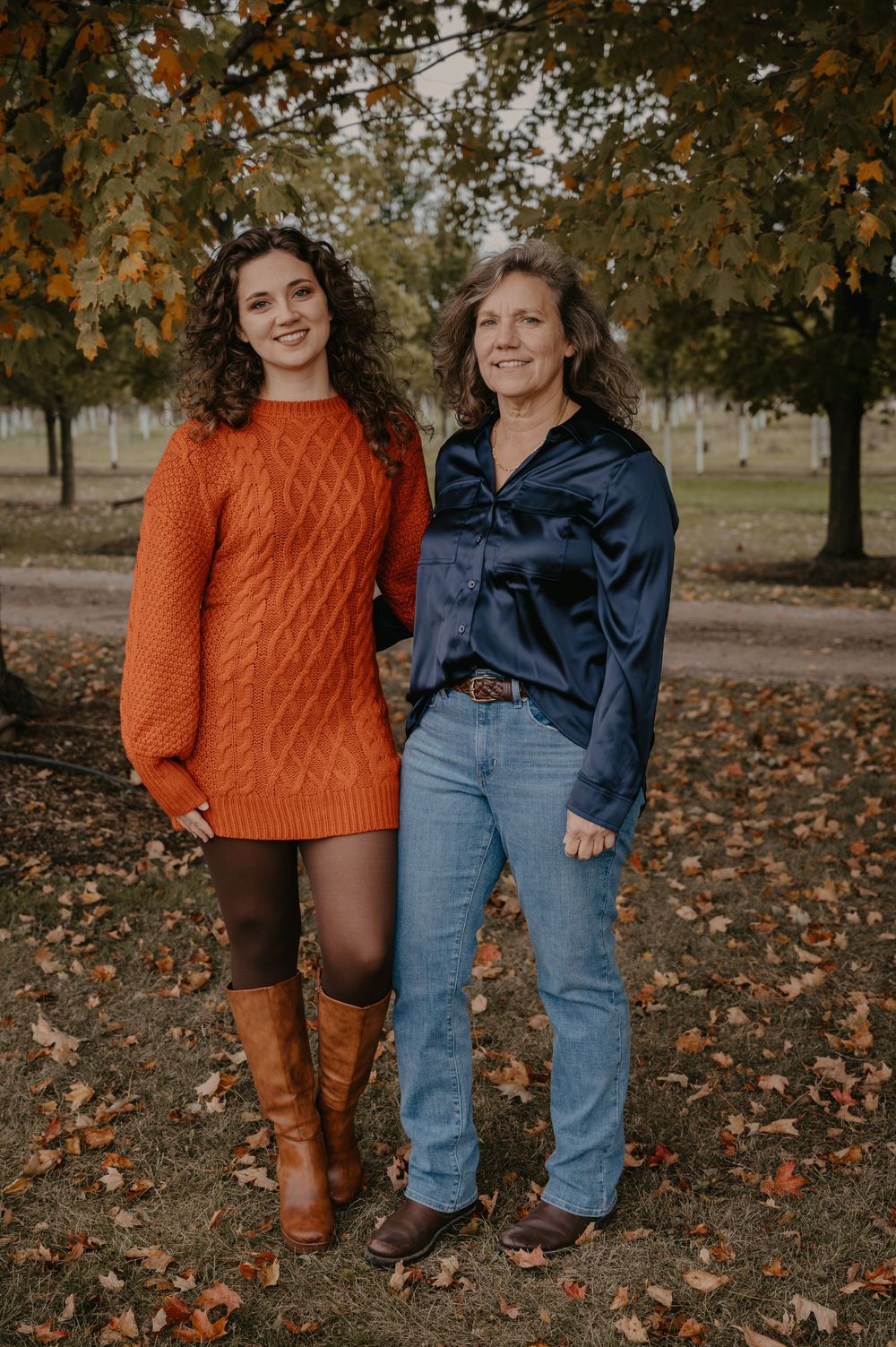 Two women smile together outdoors in fall, one in an orange knit dress with brown boots, the other in a navy blouse and jeans.