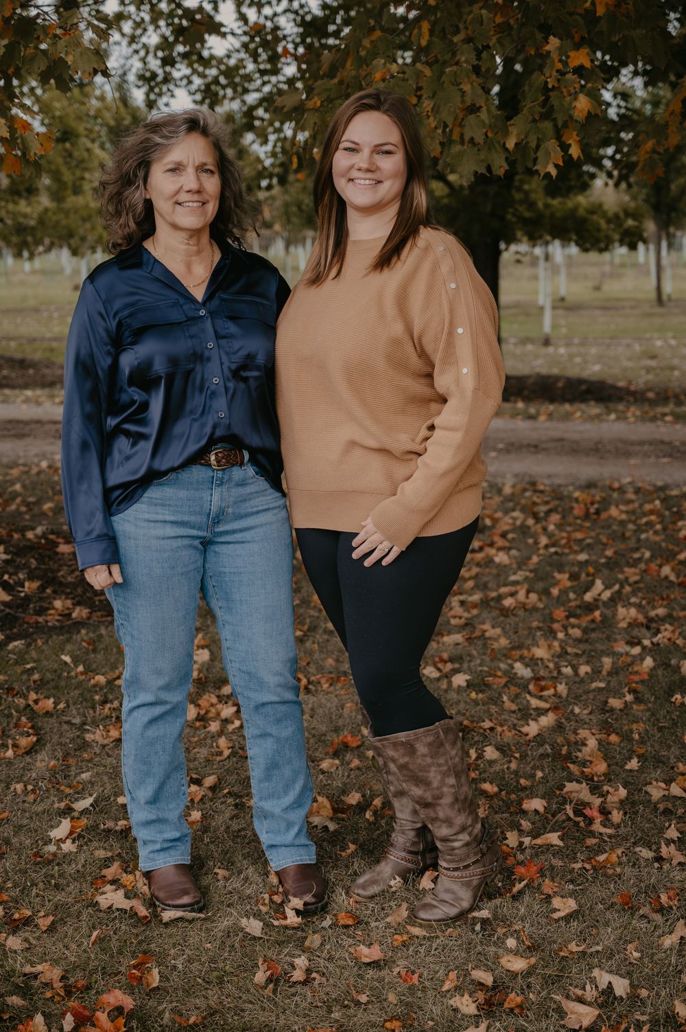 Two women stand smiling in an autumn park, one in a navy satin blouse with jeans, the other in a tan sweater with dark leggings.