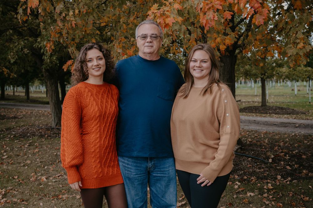 Three people smile together outdoors in fall, with a man in blue flanked by two women in orange and tan sweaters.
