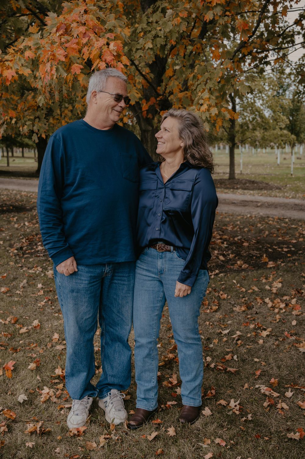 A couple smiles at each other lovingly while standing together in a park surrounded by autumn leaves.