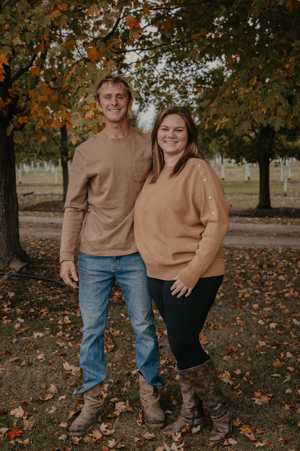 A smiling couple poses together outdoors in autumn, both wearing coordinating tan and camel toned outfits.