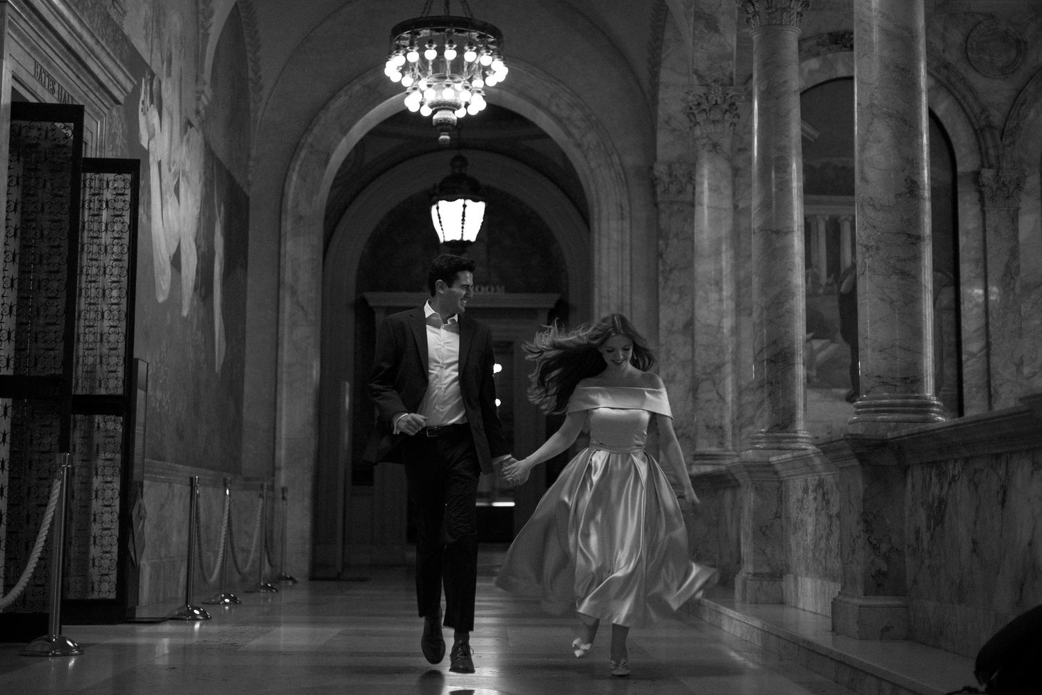 A couple dances elegantly through an ornate hallway with chandeliers and stone arches in black and white.